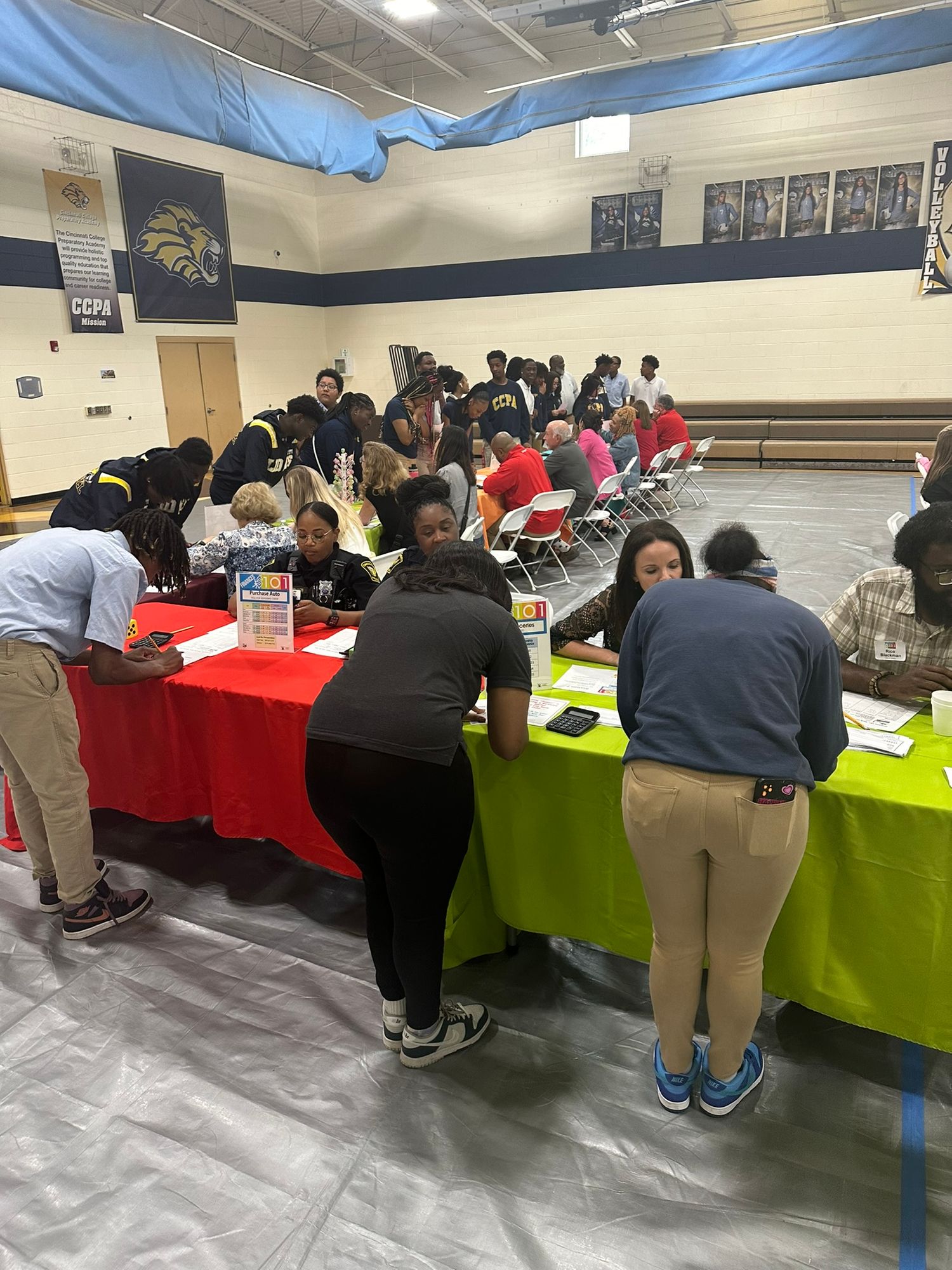 Students and staff gather around colorful tables in a school gymnasium during a busy registration or resource fair event.