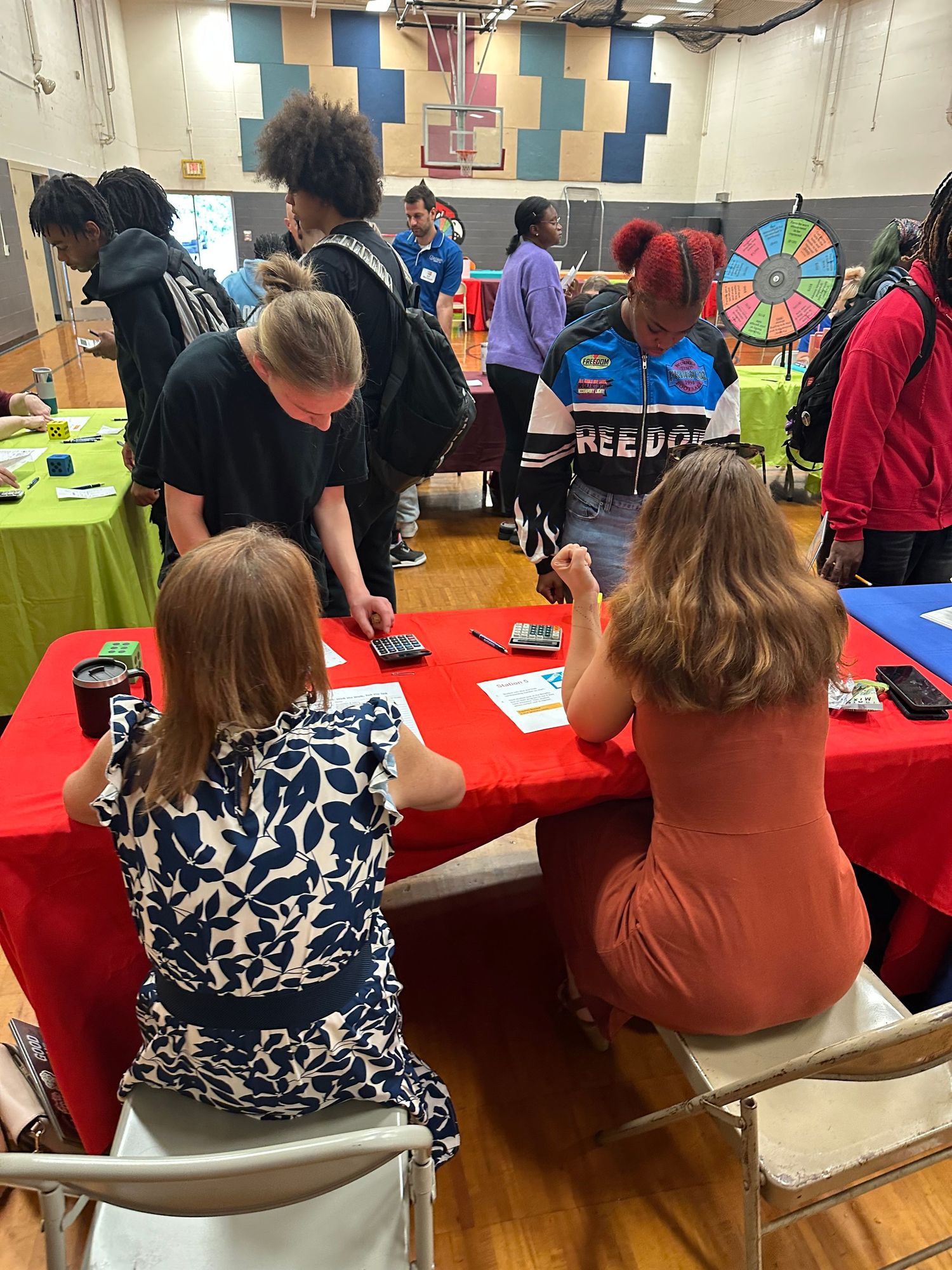 Students and educators engage at interactive tables during a school financial literacy fair in a gymnasium.