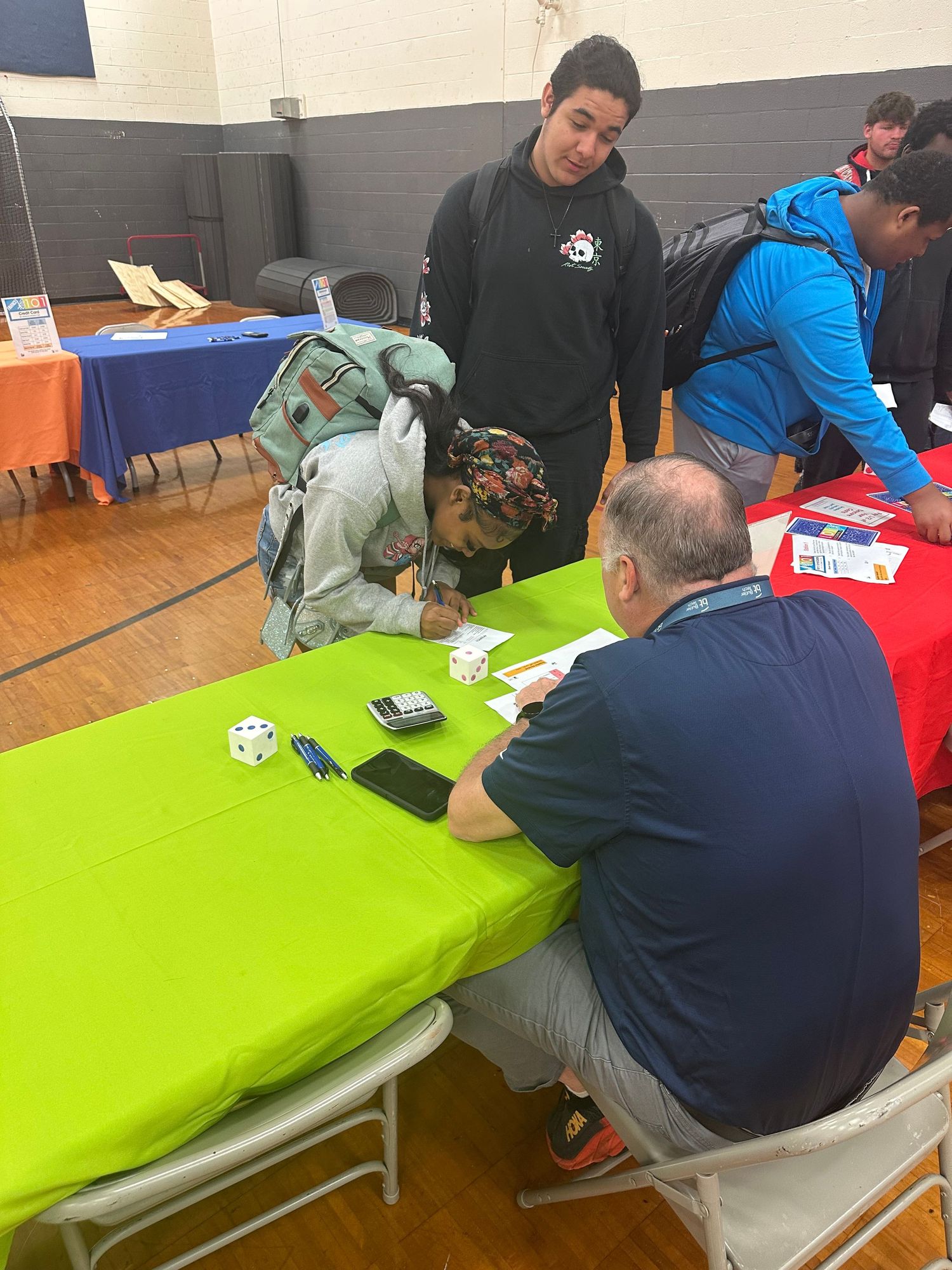 Students gather around a green table with an adult mentor at an indoor school fair event in a gymnasium.