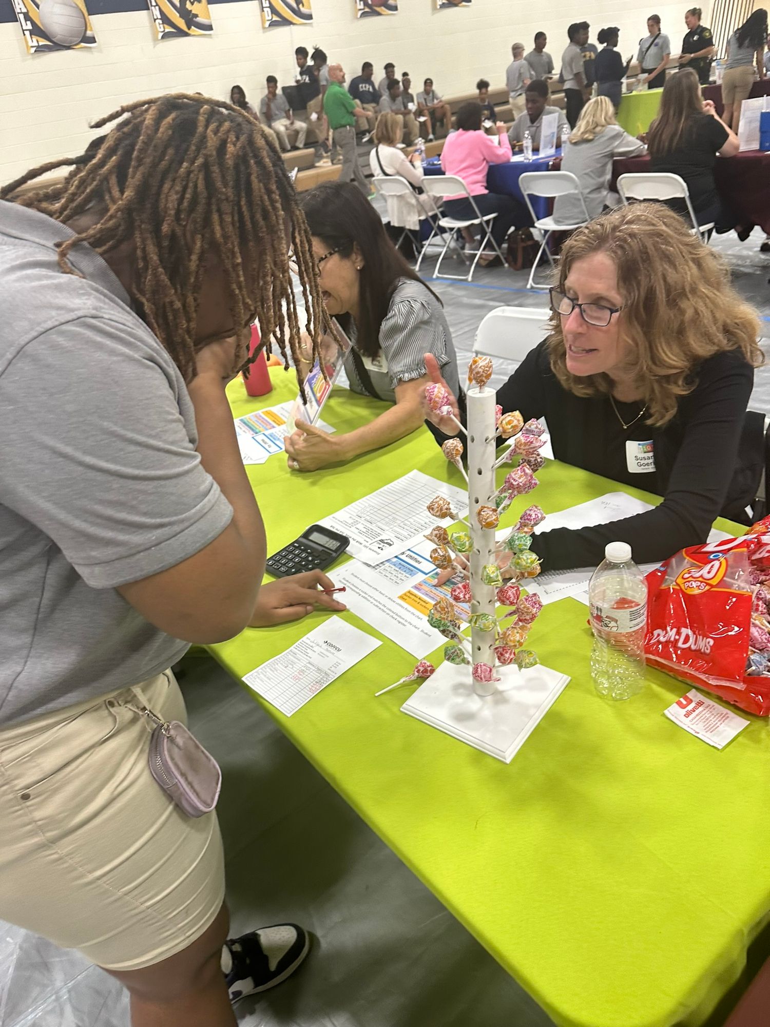 A woman assists students with paperwork at a green table decorated with a lollipop tree at a school fair.