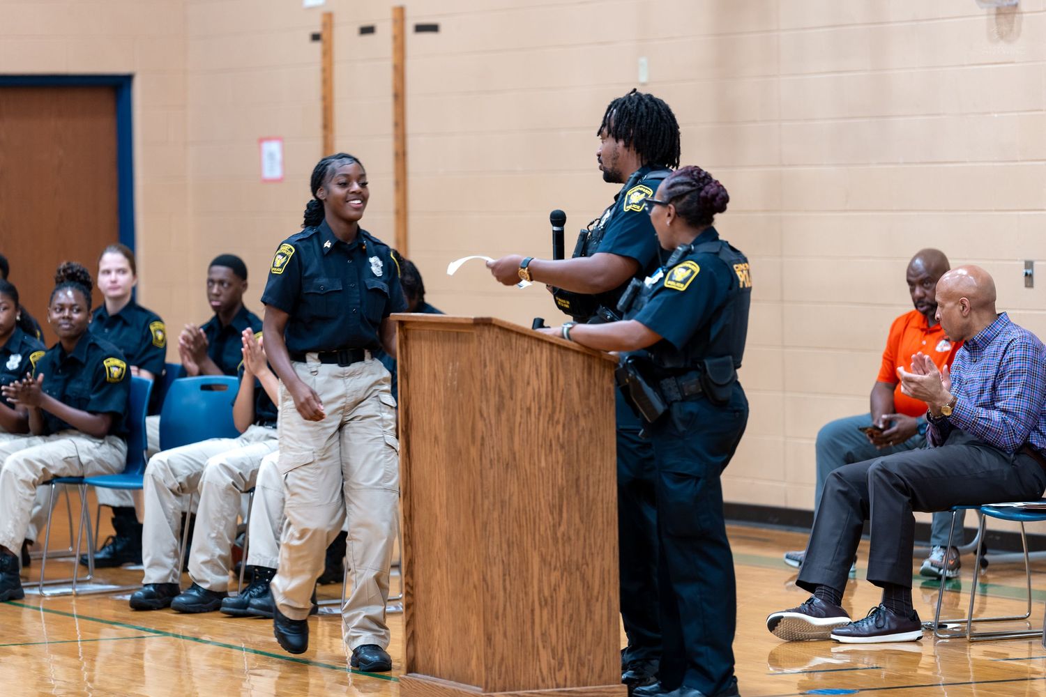 Female police officers stand at a podium during an awards ceremony in a school gymnasium with seated officers nearby.