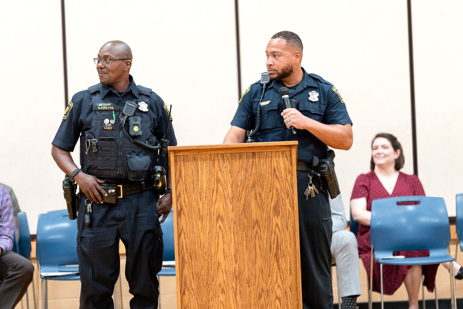 Two police officers stand at a podium speaking at a community meeting in a conference room.
