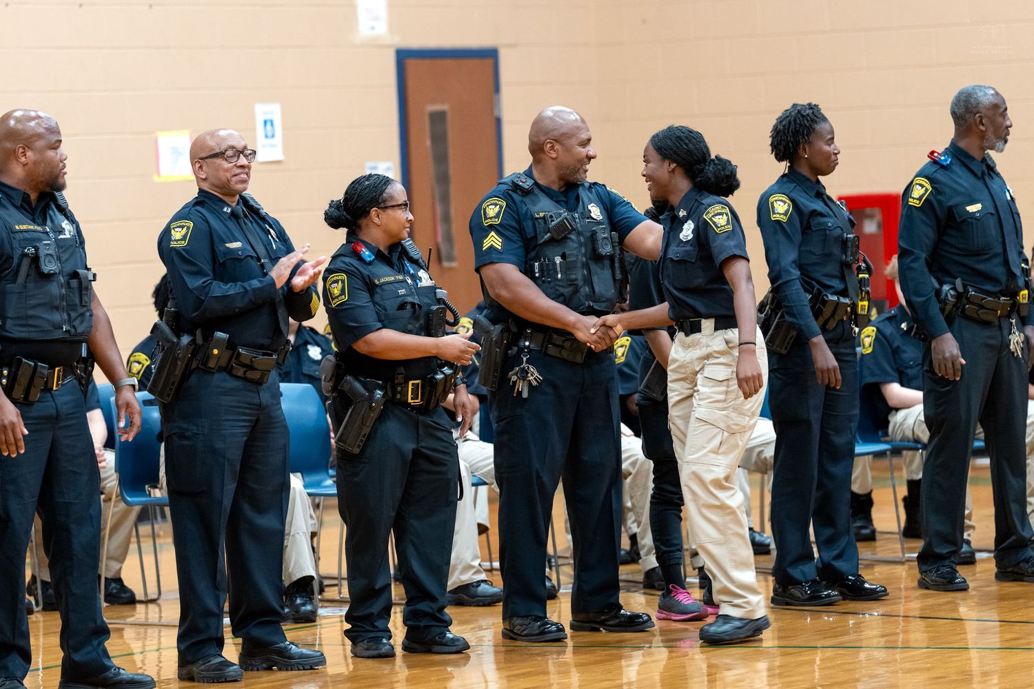 Black police officers in uniform stand in a line during an indoor ceremony in a gymnasium.