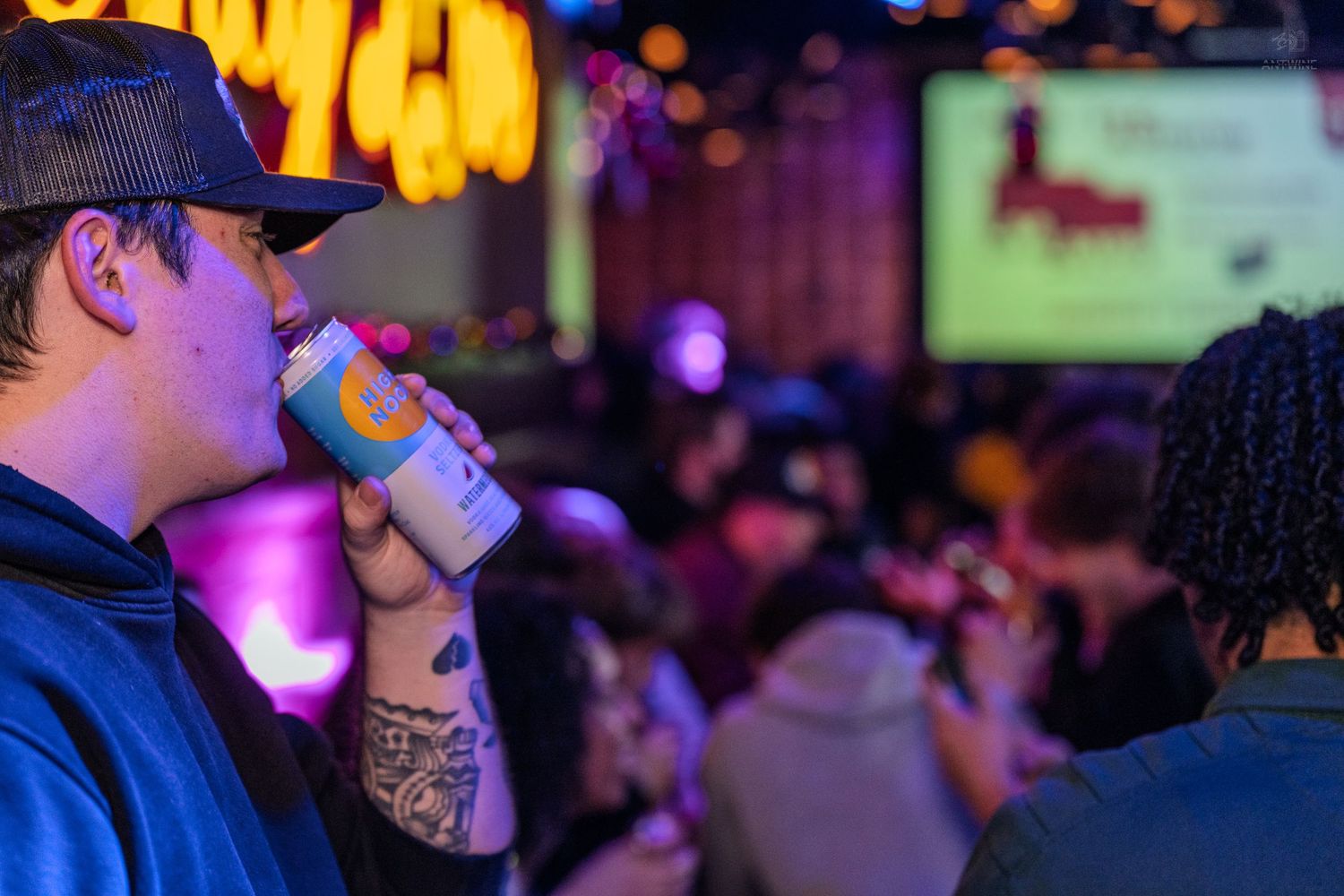 Tattooed man drinking a canned beverage at a colorful, crowded bar with neon lights.