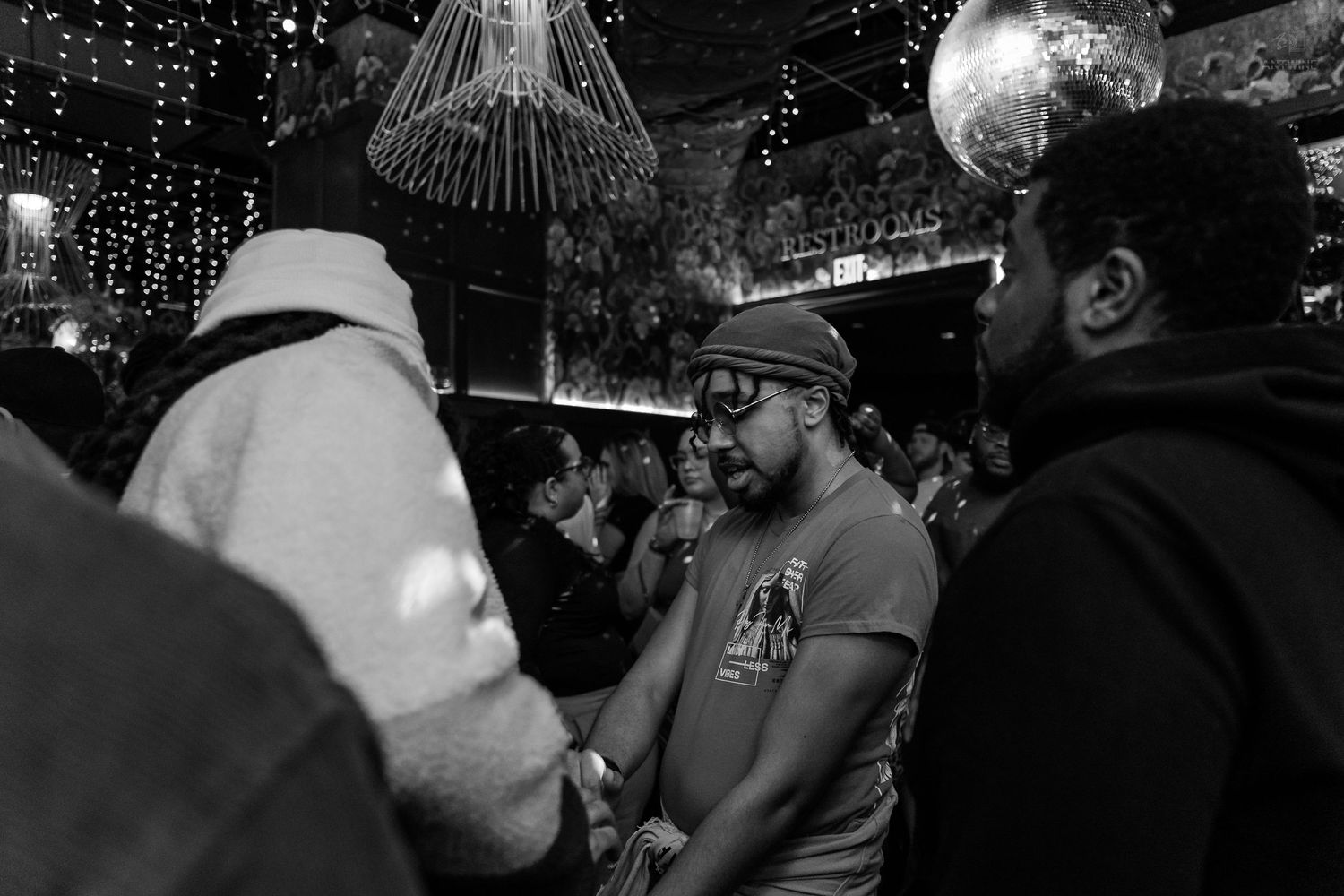 Black and white photo of men conversing in a crowded nightclub with disco ball and string lights above.