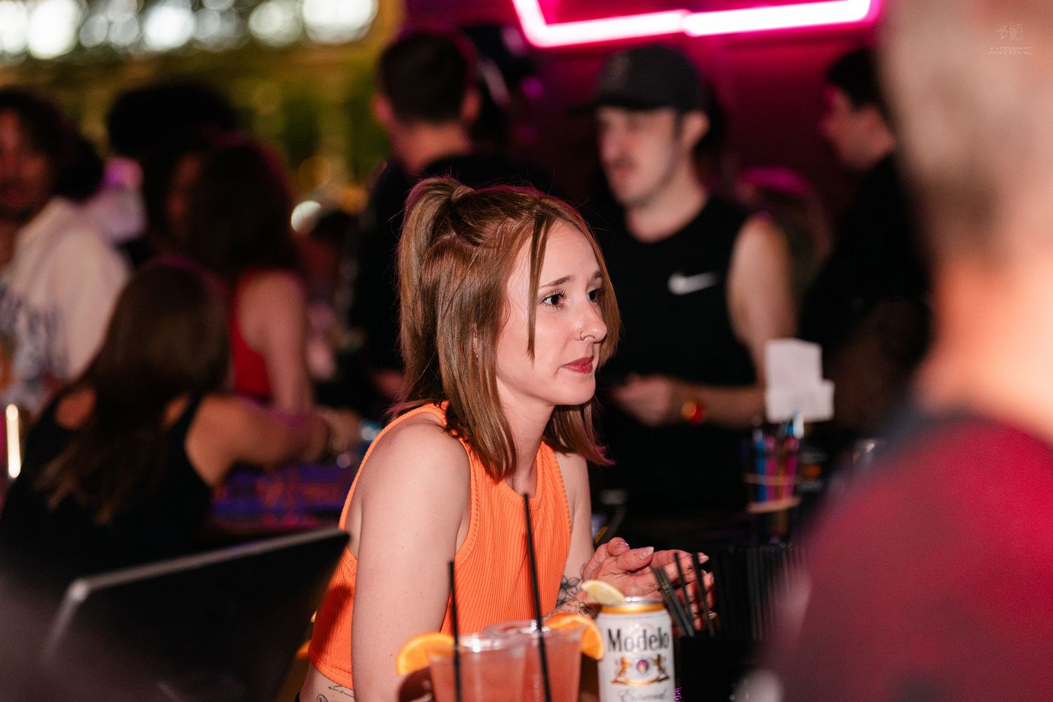 Young woman in orange top holding a drink at a crowded bar with colorful neon lights in background.