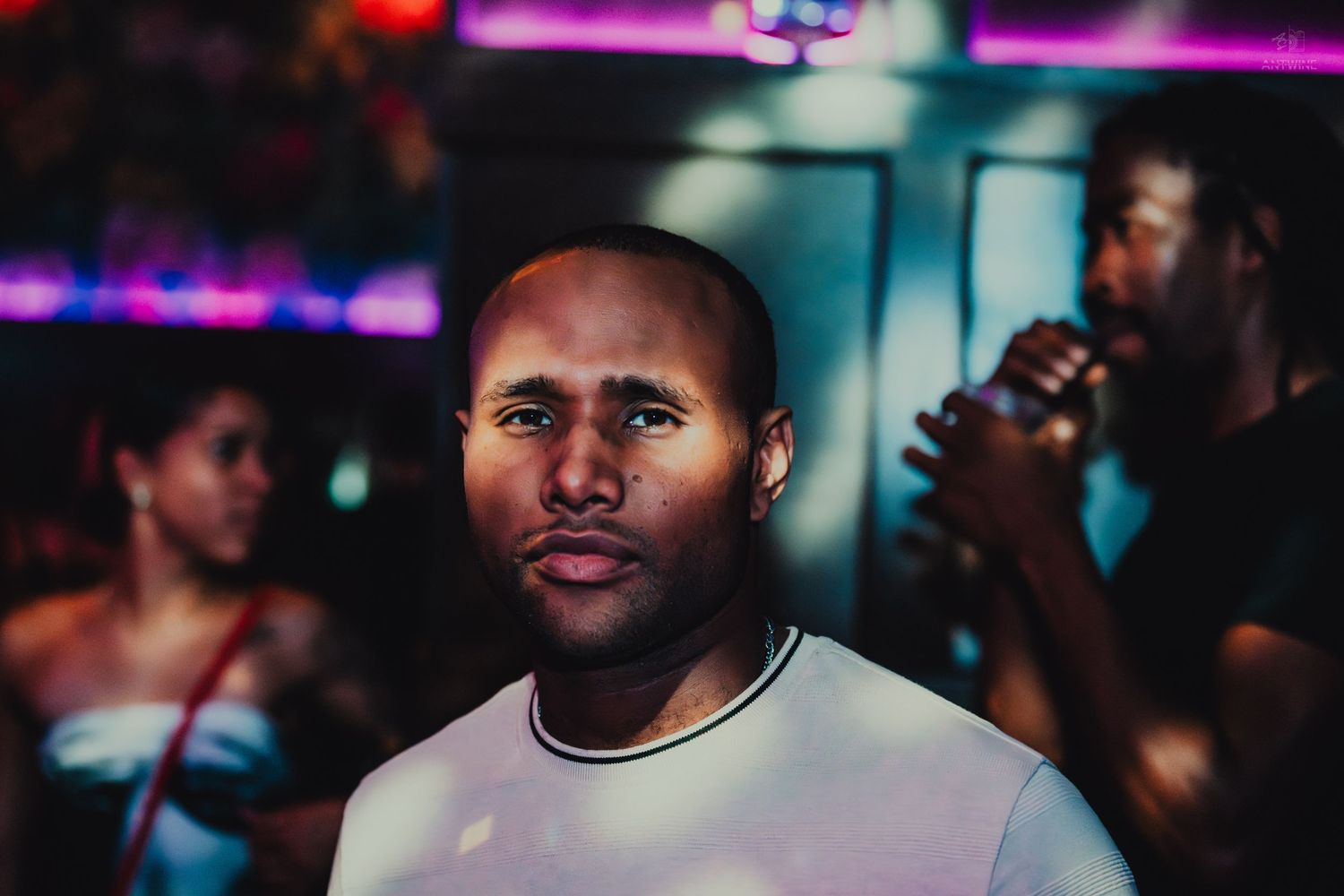 A bald Black man in a white t-shirt stands in a colorful nightclub with people in the background.