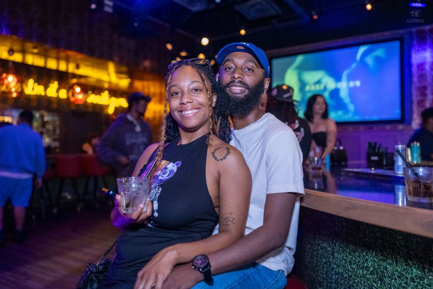 A couple smiles together at a bar, with colorful neon lighting and a screen in the background.