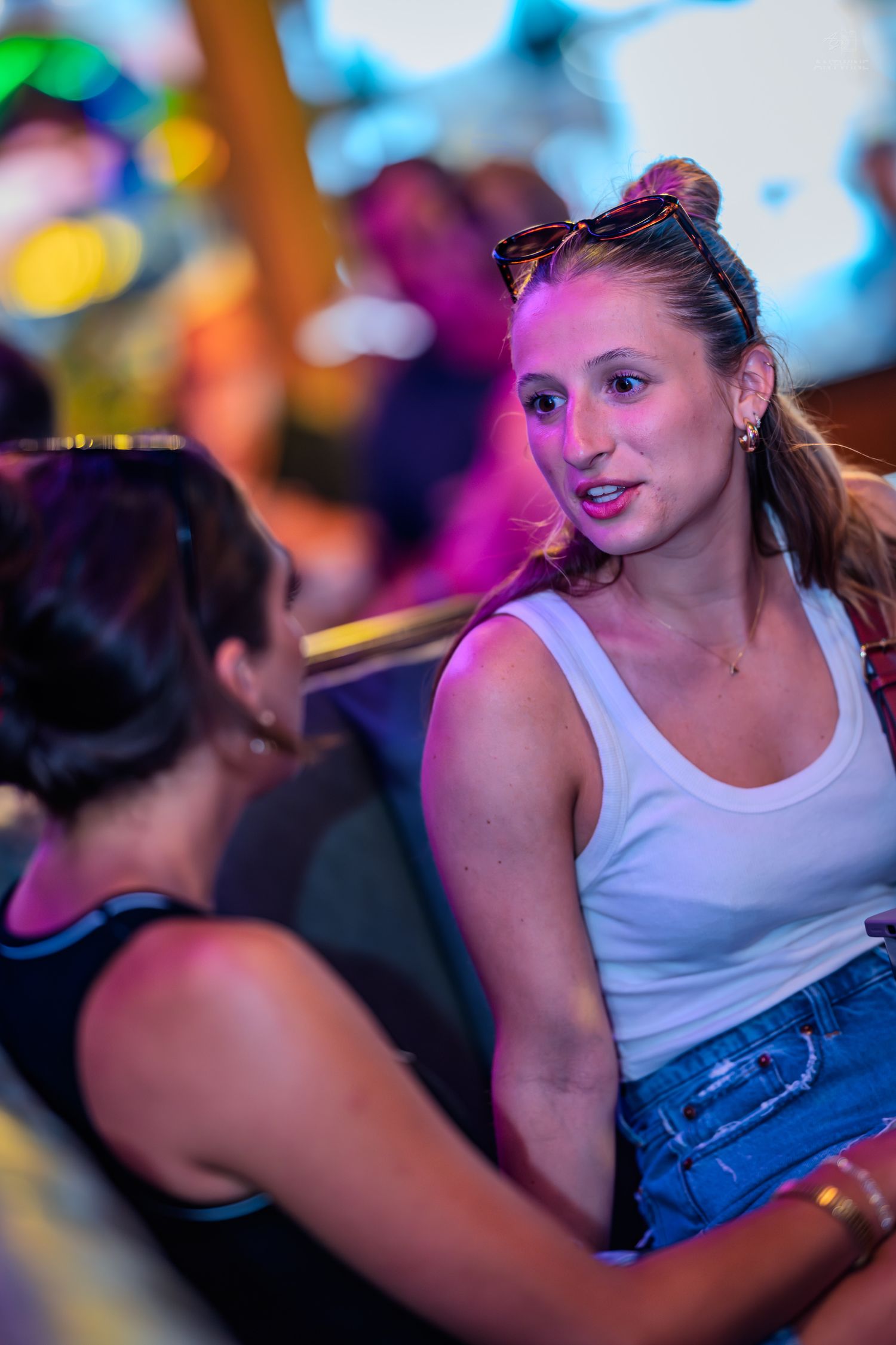 Two women chat at a vibrant nightclub, one in a white tank top and denim shorts.