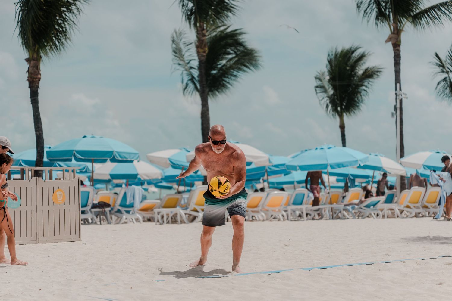 Shirtless man playing paddleball on a tropical beach with colorful blue umbrellas and lounge chairs behind him.