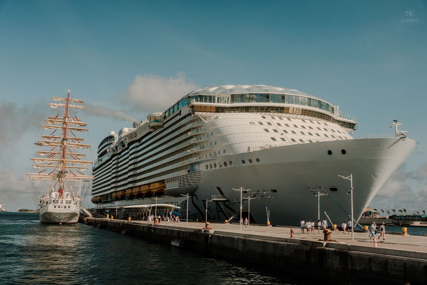 A massive Royal Caribbean cruise ship docked at a sunny Caribbean port next to a tall sailing vessel.