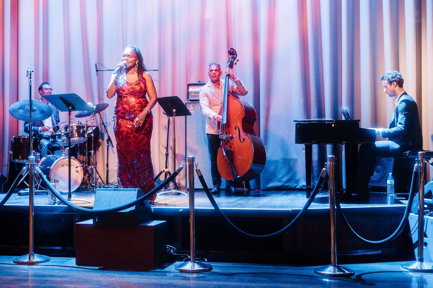 Female singer in red sequin gown performs with jazz band including upright bass and piano.