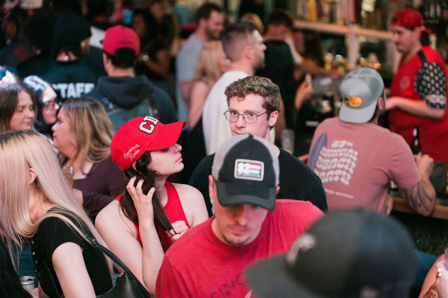 Young adults in baseball caps mingle at a packed Cincinnati bar event with staff nearby.