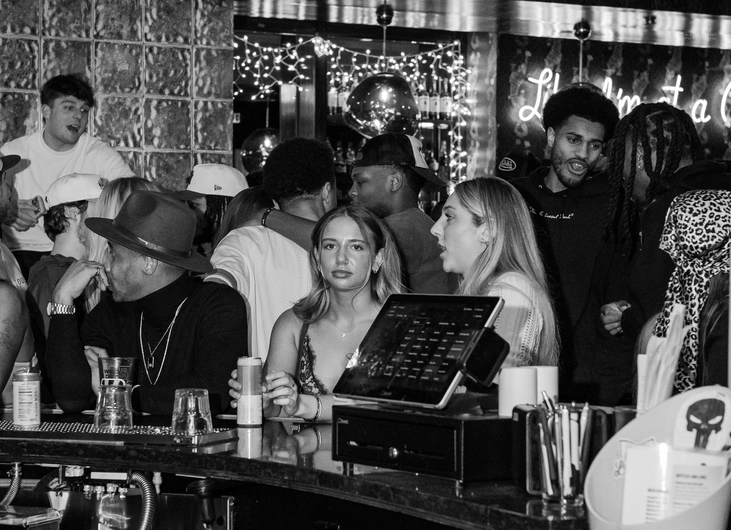 Black and white photo of bar patrons socializing near a POS terminal at a nightclub.