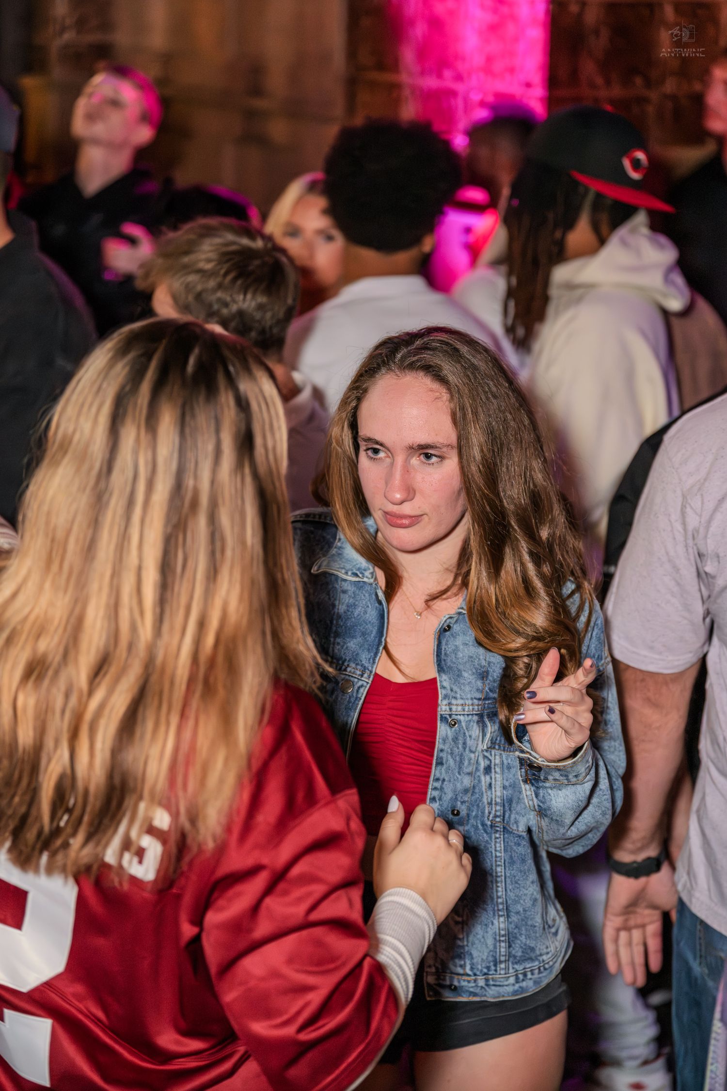 Young woman in denim jacket and red top looking at camera at a crowded nightclub.