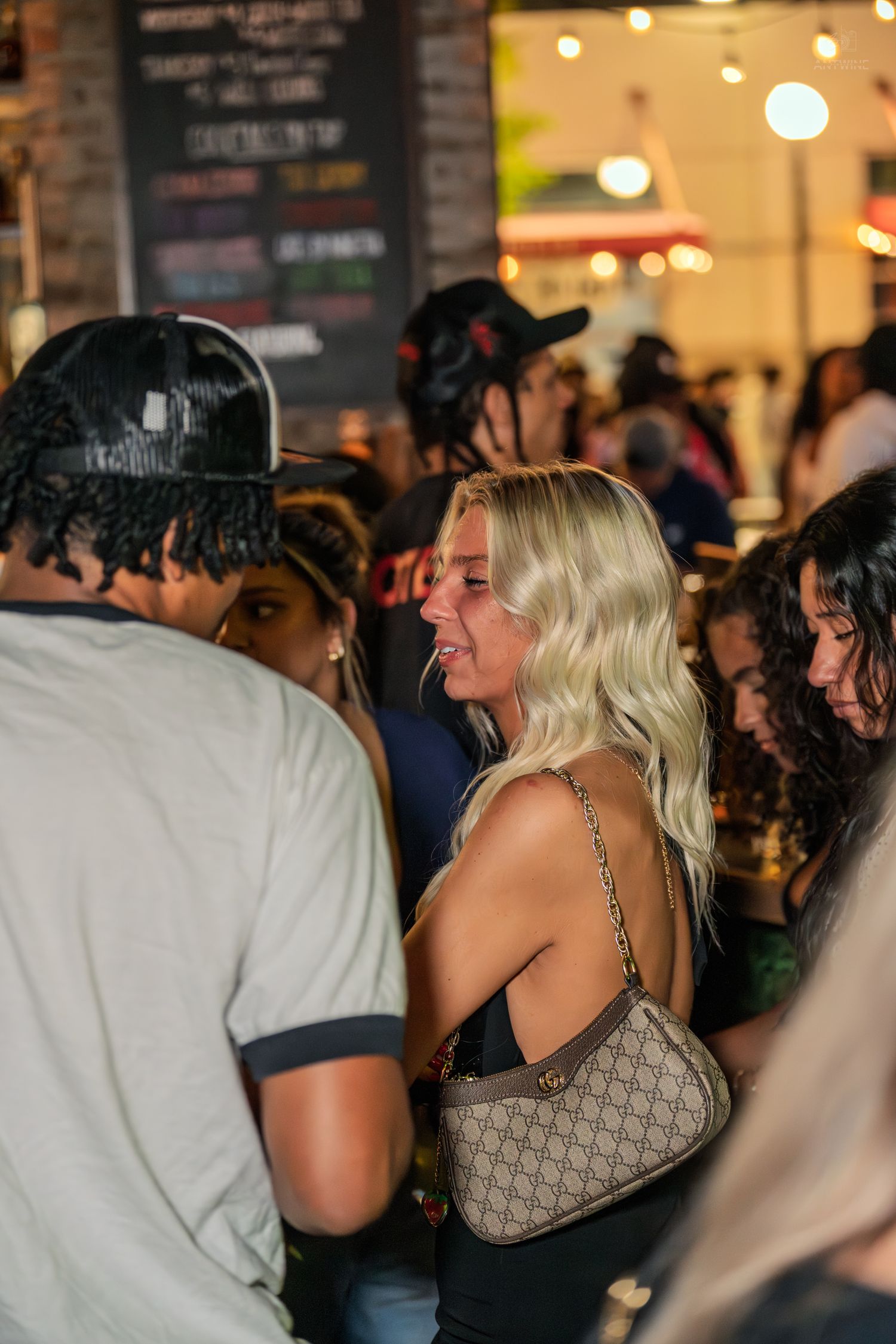 Blonde woman in a black top carrying a Gucci bag at an outdoor bar gathering.