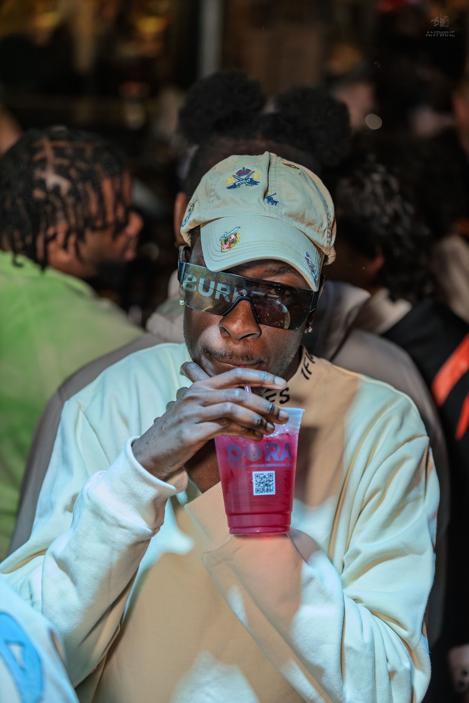 Man in beige cap and sunglasses sipping pink drink at party.