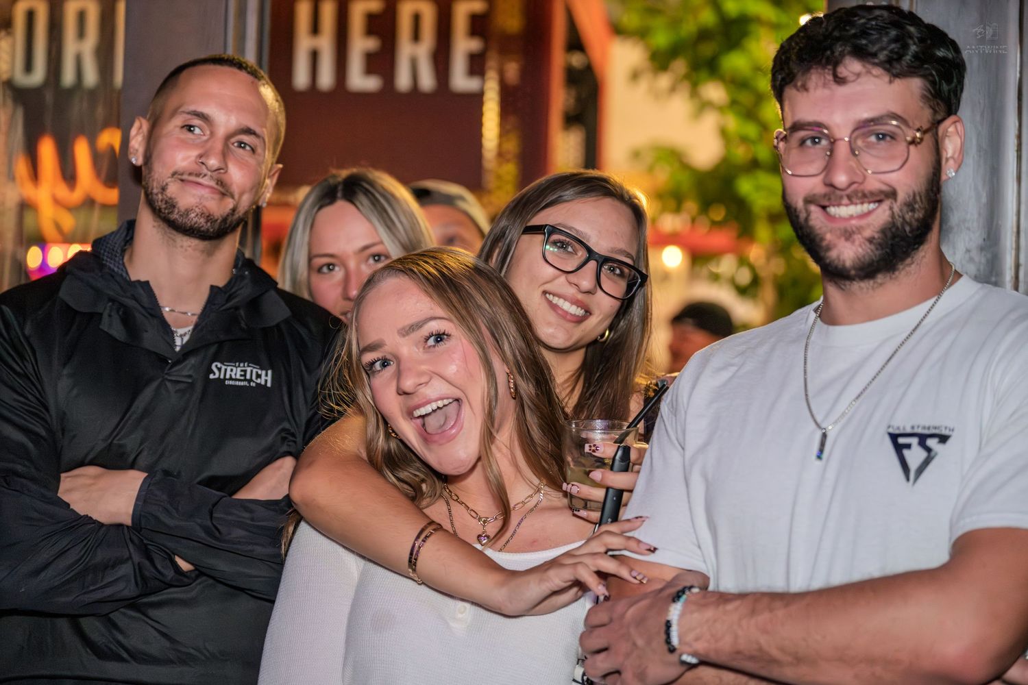 Group of five young adults laughing and posing outside a bar at night.