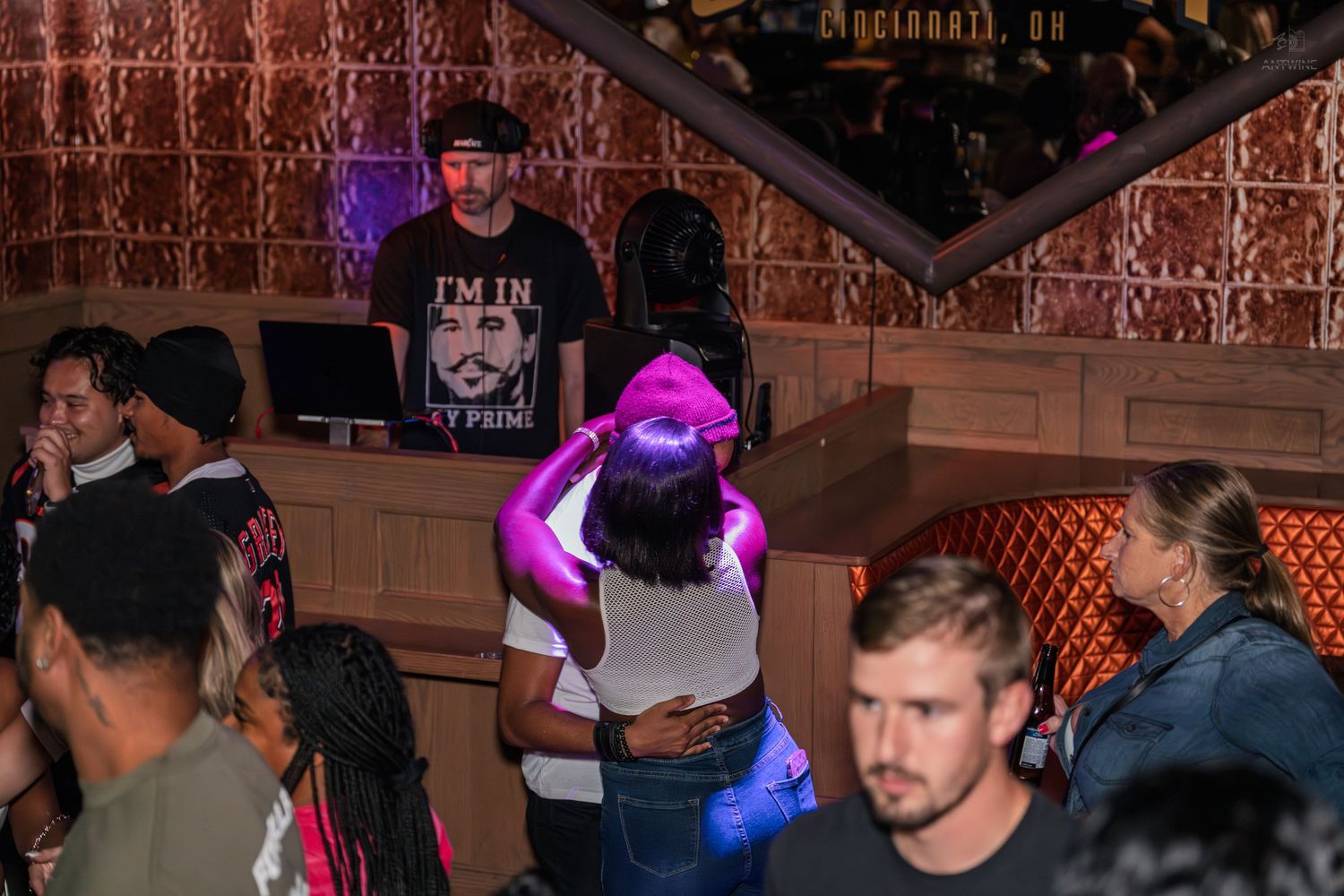 Crowd dancing under purple lights at a nightclub with a DJ in the background.
