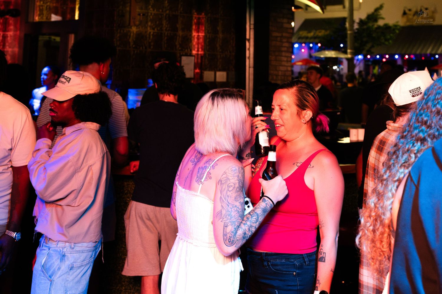 Two women chatting at an outdoor bar, one with tattoos holding a beer bottle at a nighttime event.