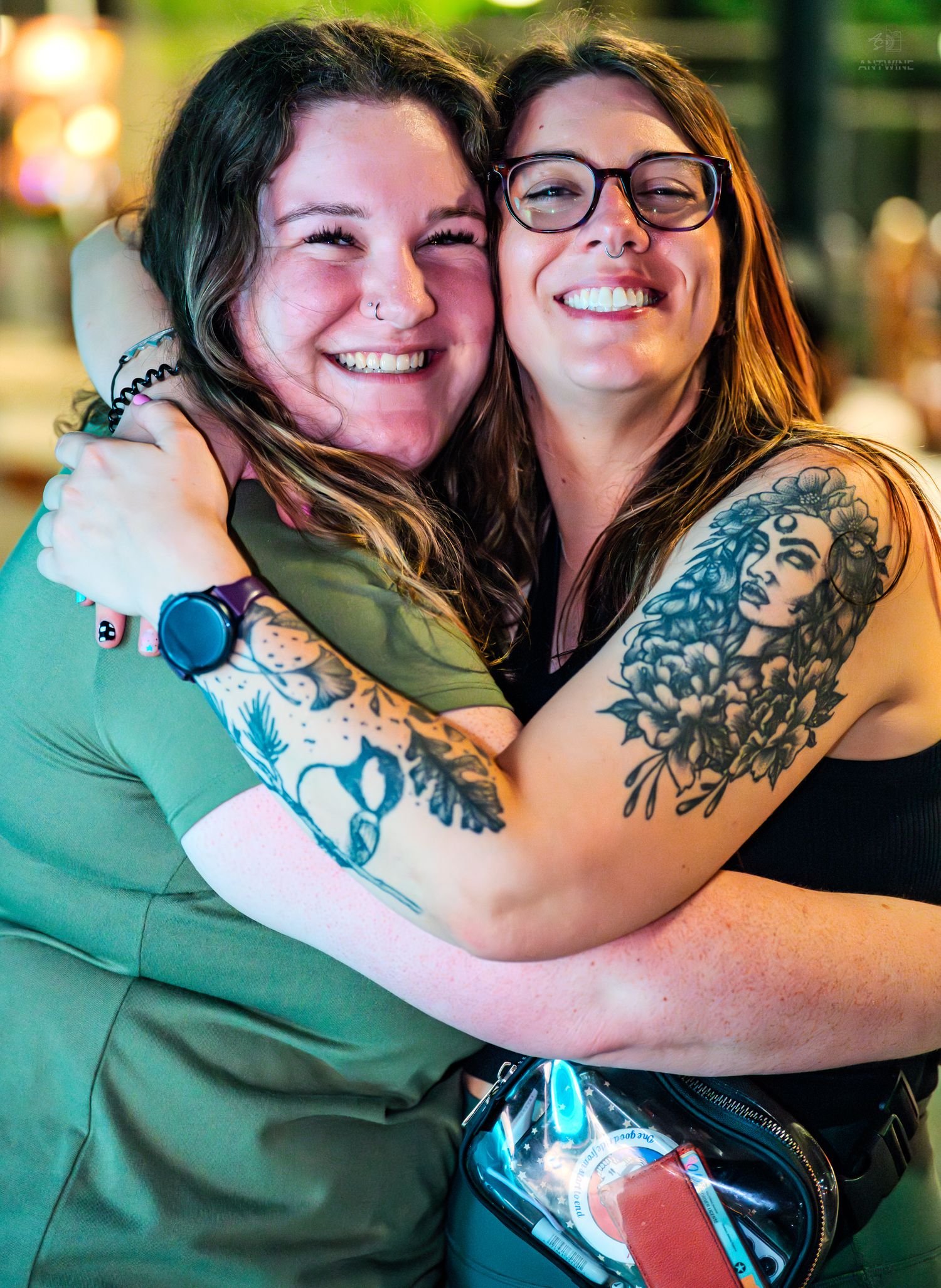 Two happy women hugging outdoors, one with a large floral tattoo sleeve at a social gathering.