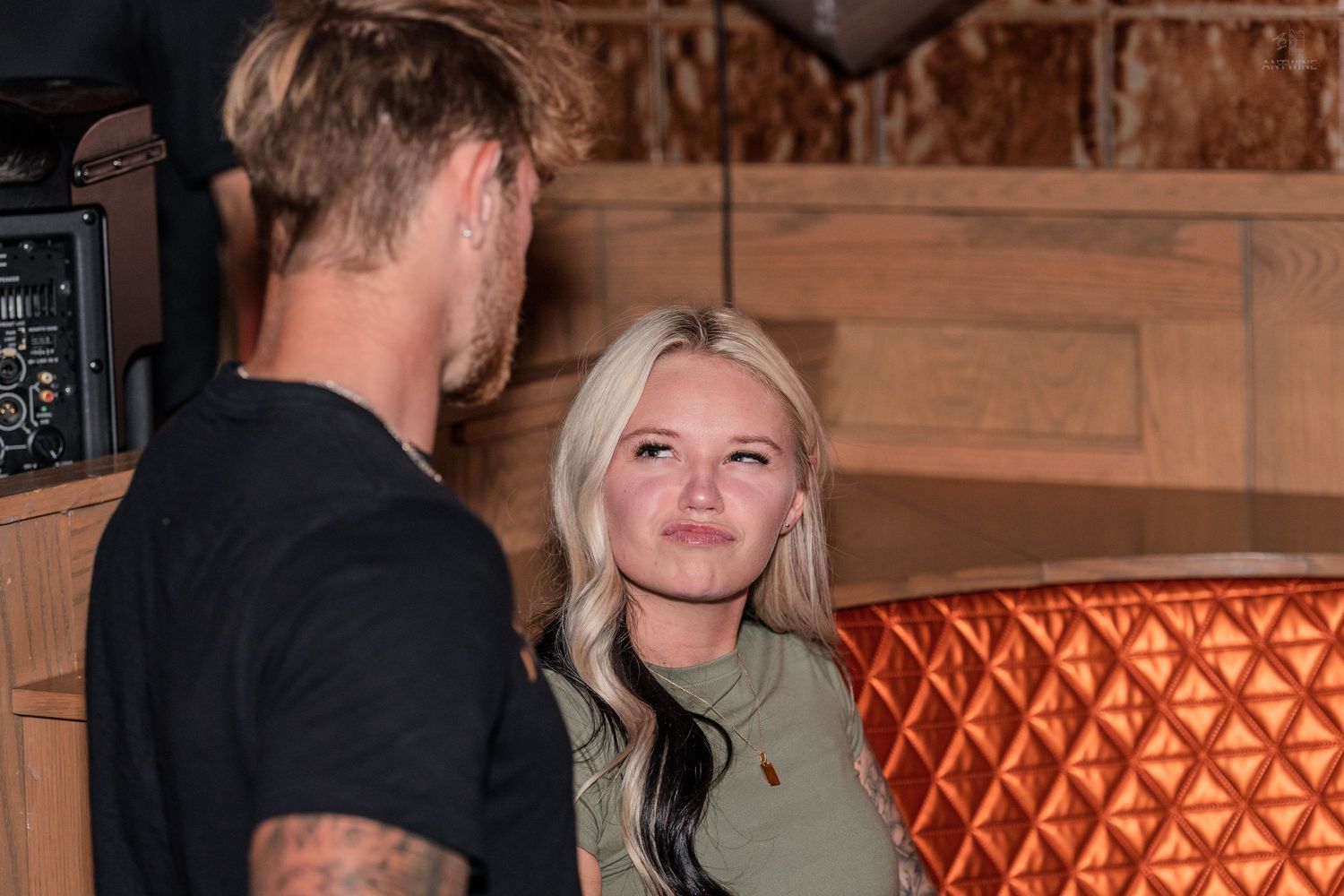 Blonde woman in green top talks with a tattooed man at a bar lounge.