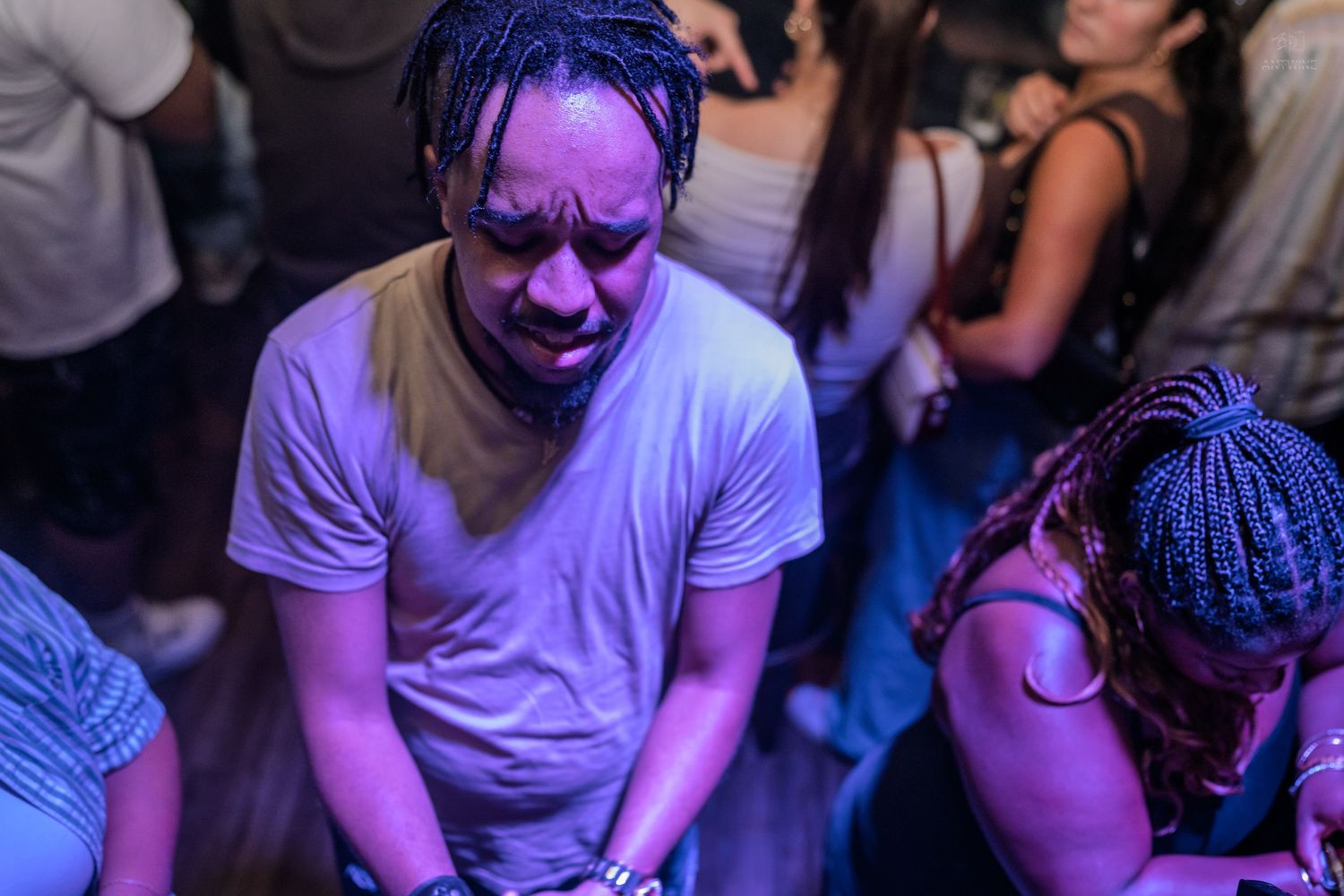 Man in white t-shirt dancing at a purple-lit nightclub with crowd around him.