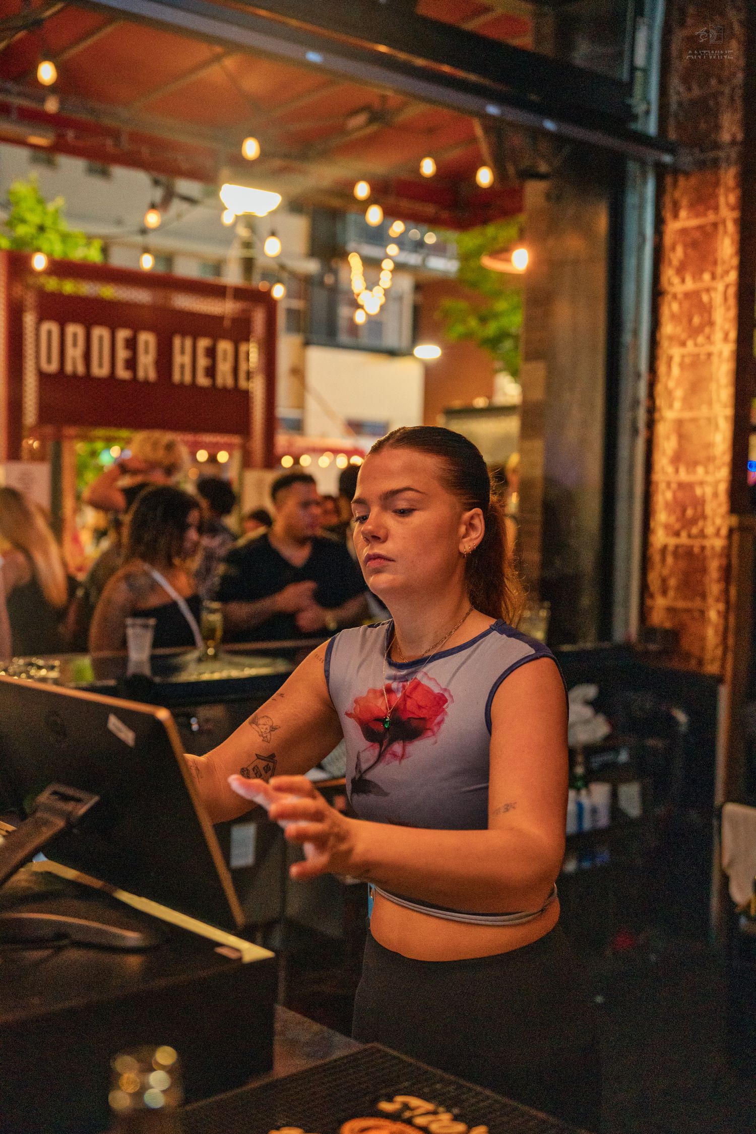 Female bartender working at a POS terminal near an Order Here sign at a lively outdoor bar.