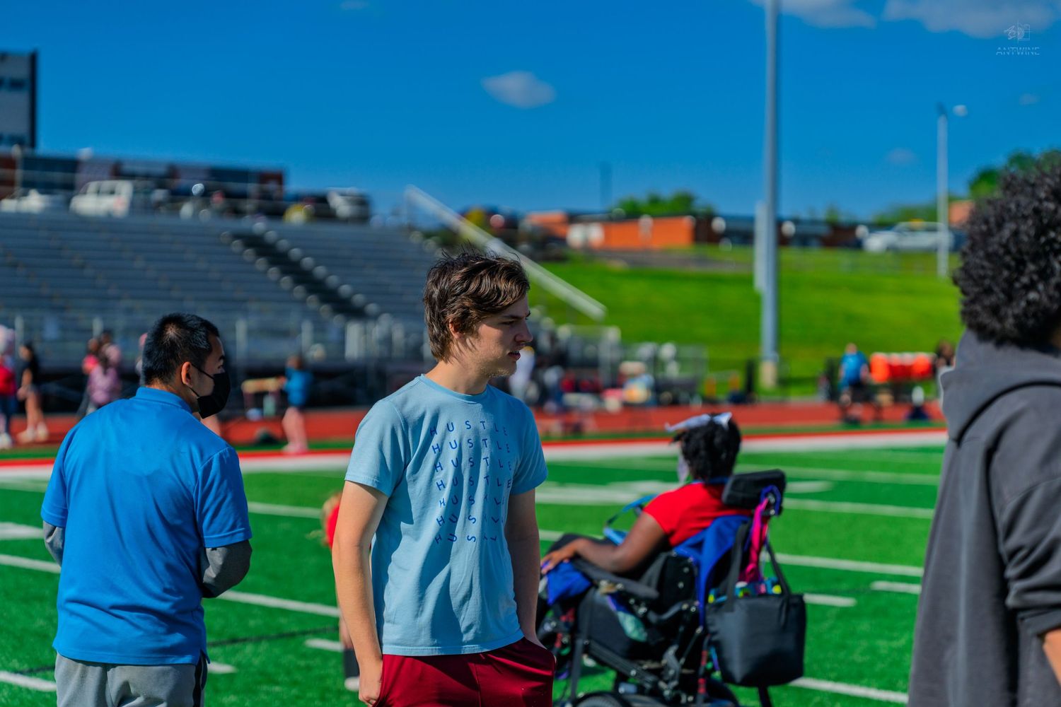 People standing near a wheelchair user on a green athletic field stadium on a sunny day.