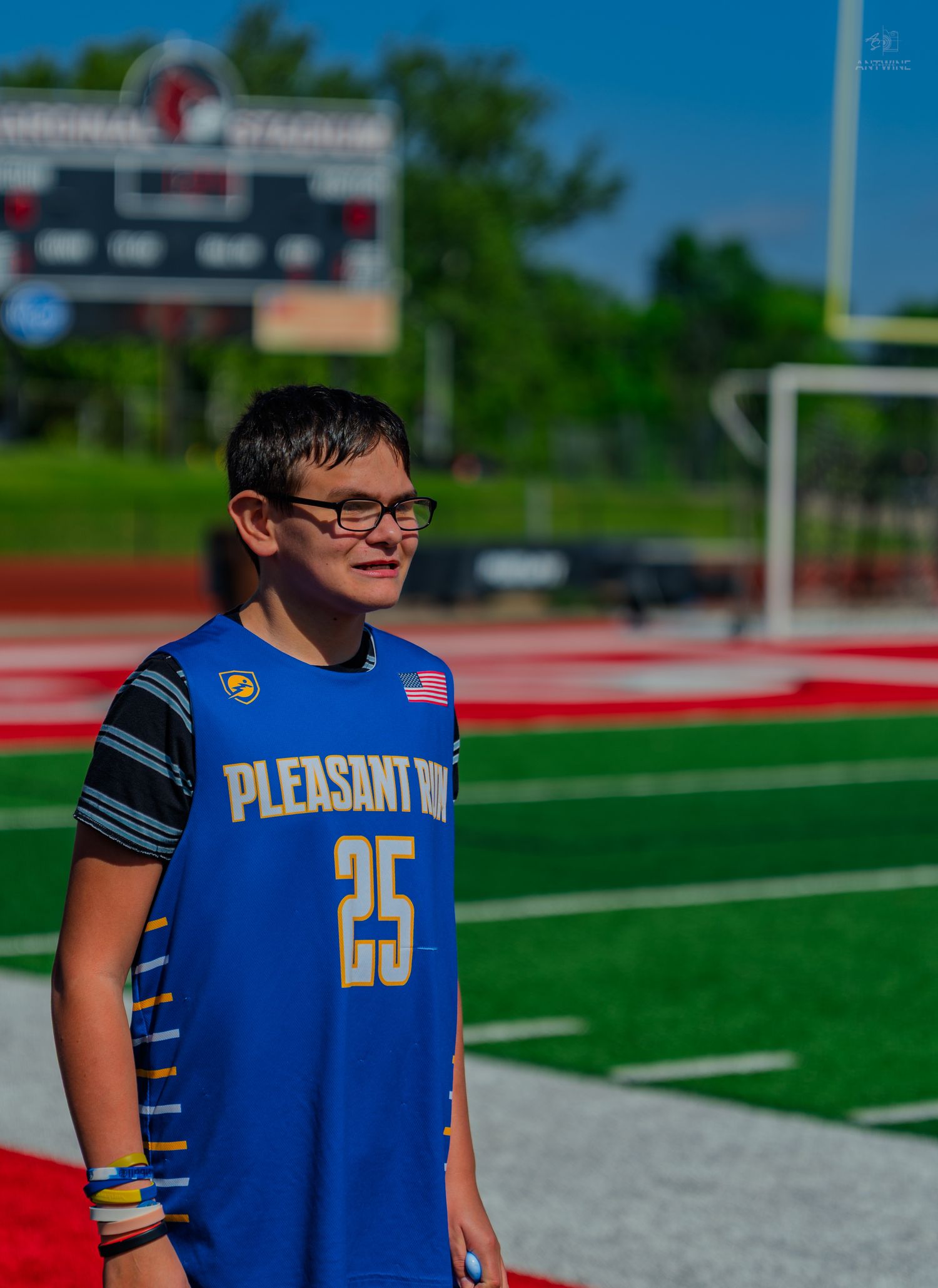 Young athlete wearing Pleasant Hill #25 blue jersey stands on red turf football field with scoreboard in background.