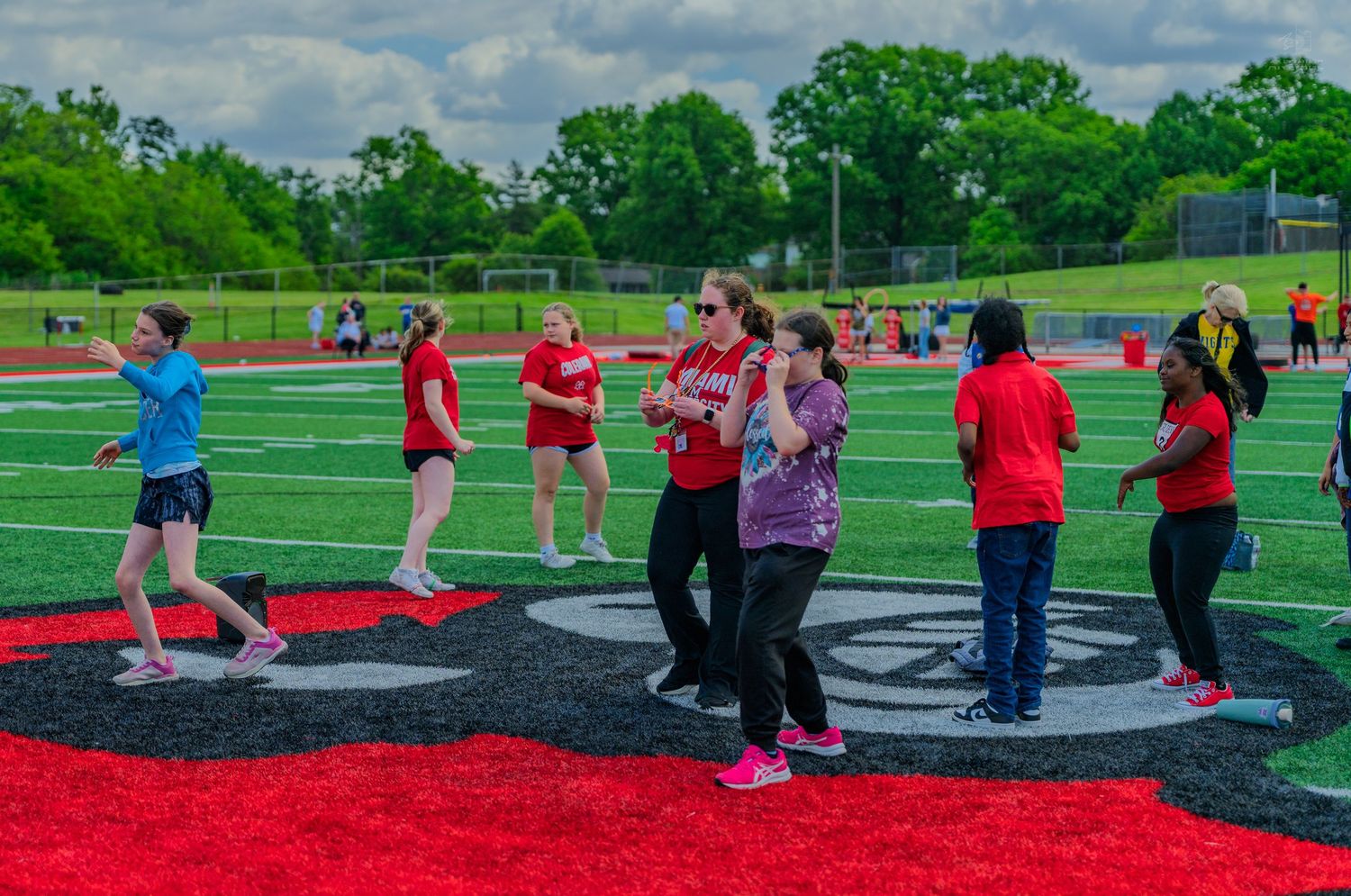 Students walk and socialize on a red and black turf field during an outdoor school event on a cloudy day.