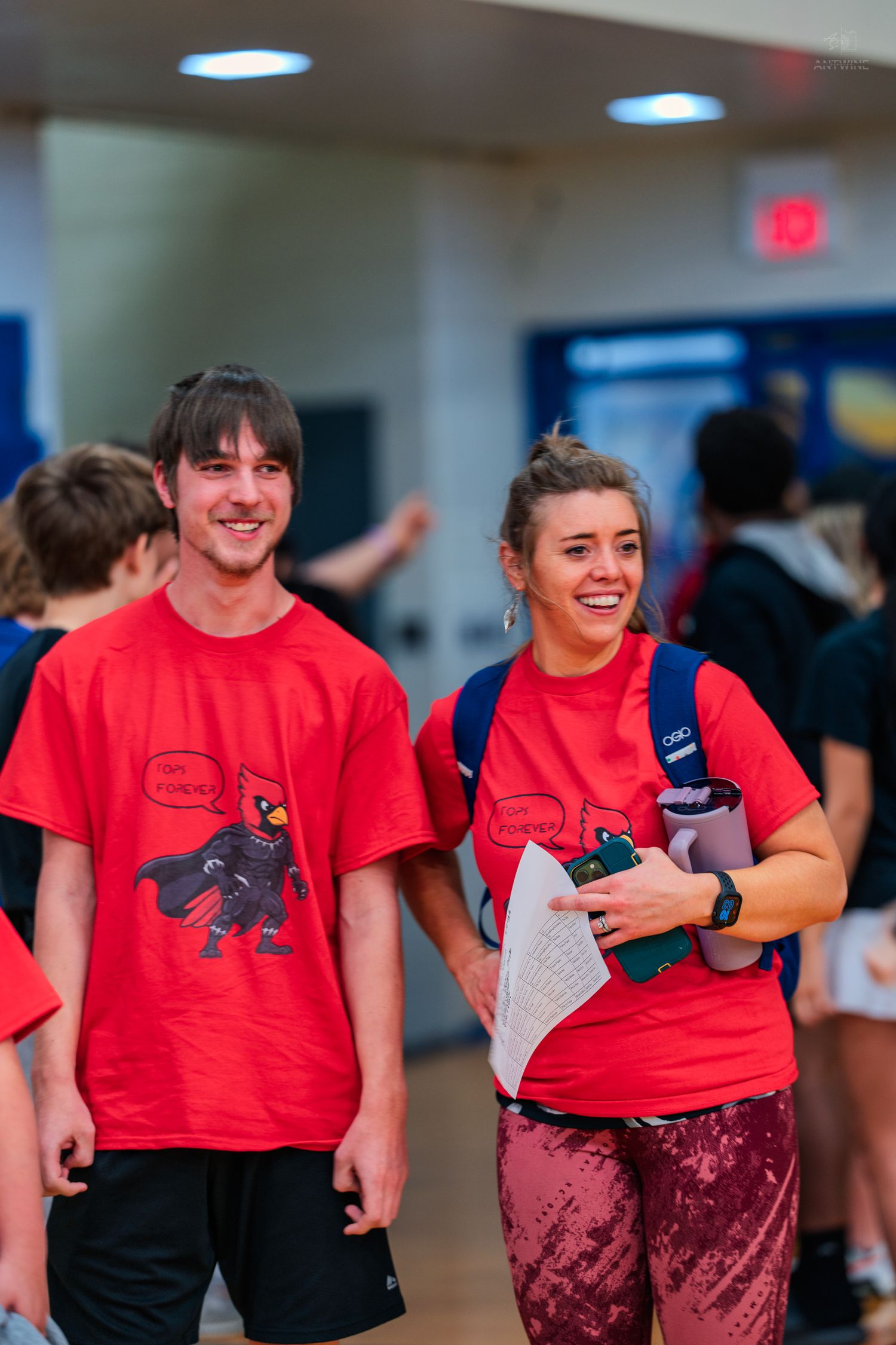 Two smiling students in matching red cardinal mascot t-shirts walk through a school hallway together.