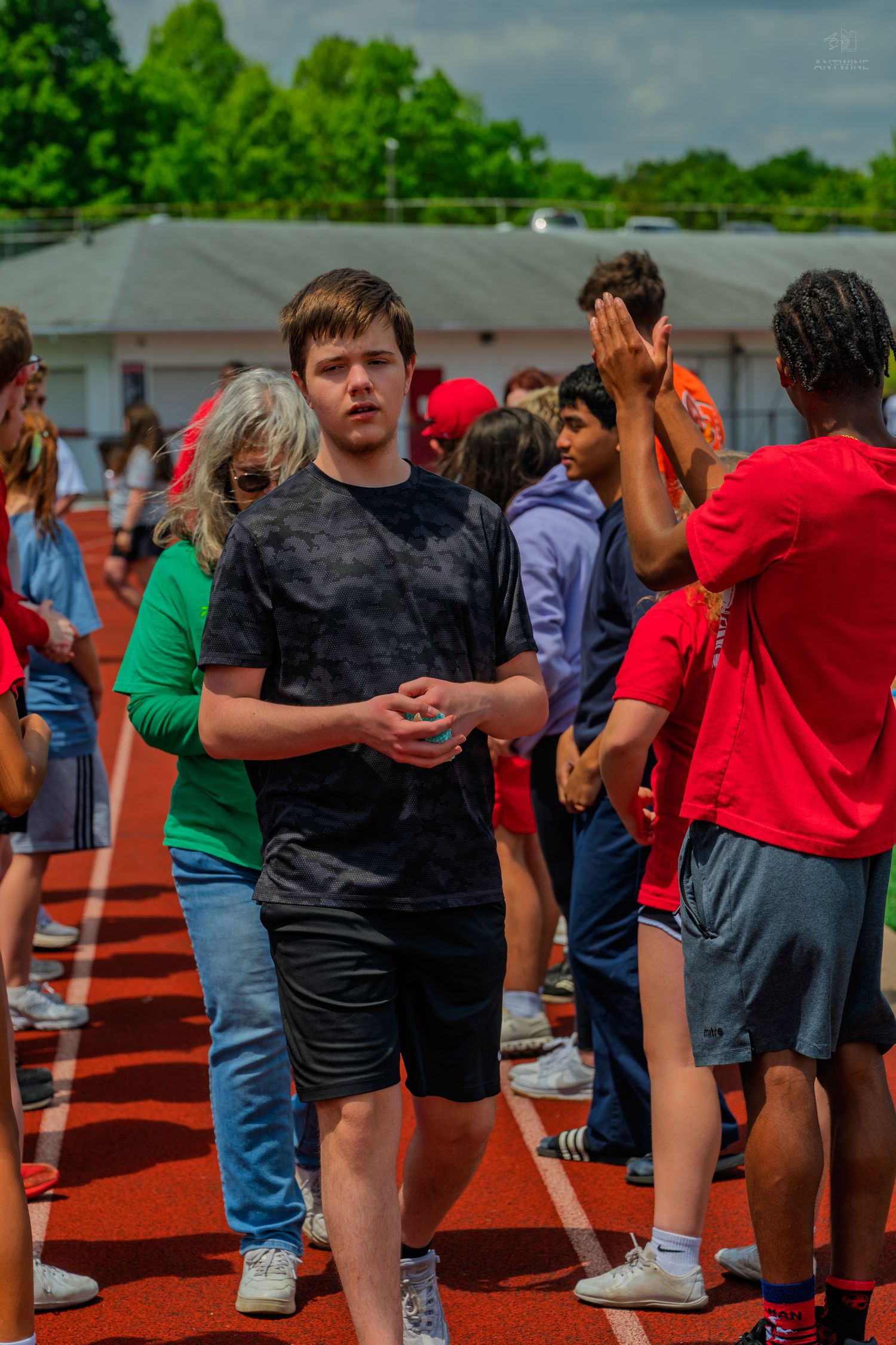 Young teen in black t-shirt and shorts walks on red track surrounded by peers at an outdoor school sports event.