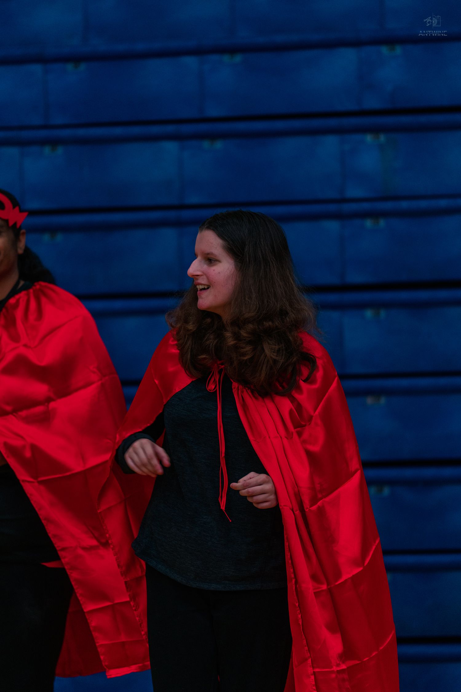 Woman wearing a red satin cape over black outfit, smiling and laughing indoors near blue bleachers.