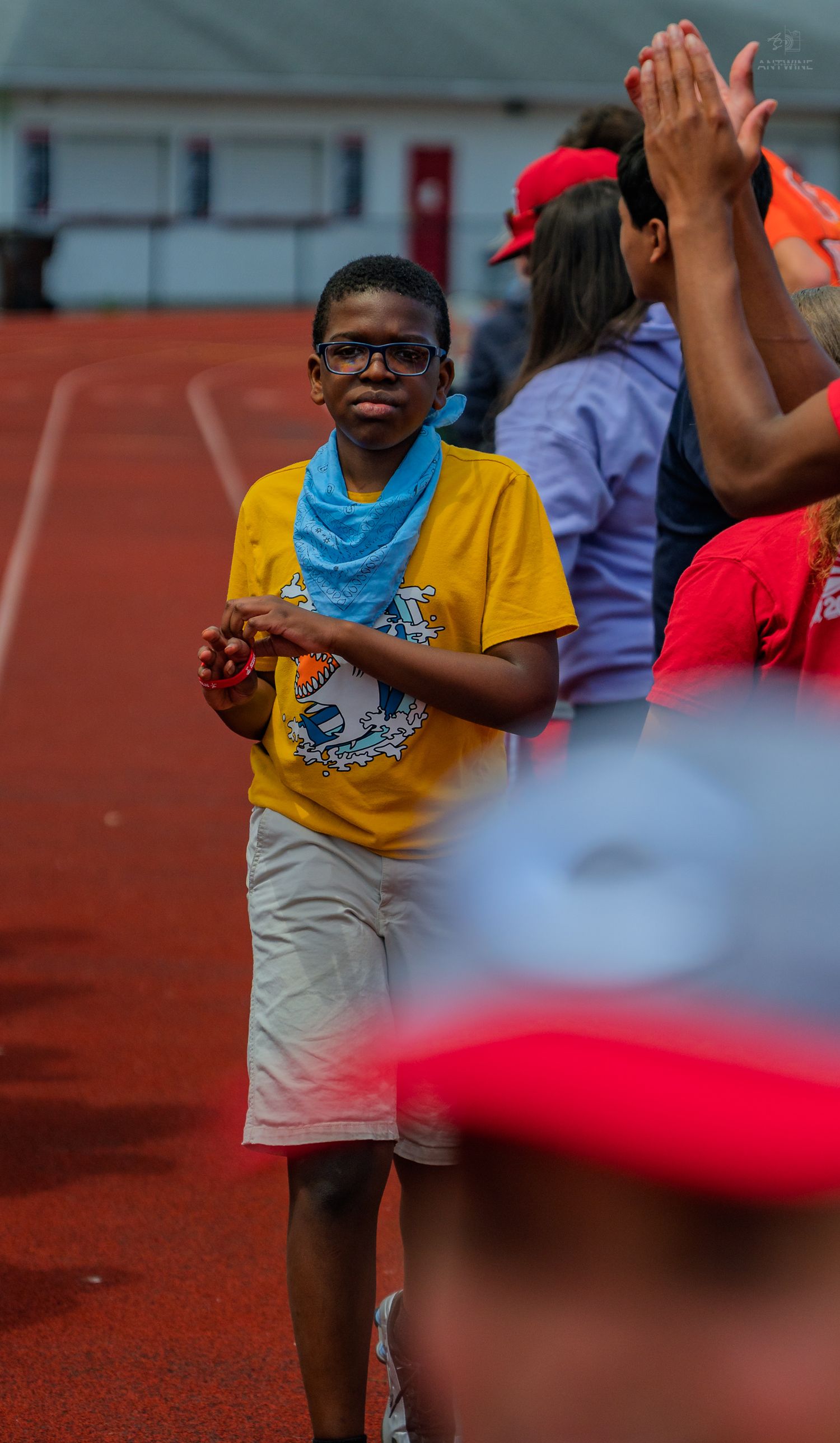 Young boy in yellow shirt and glasses walks on a red track, smiling, with people clapping in the background.