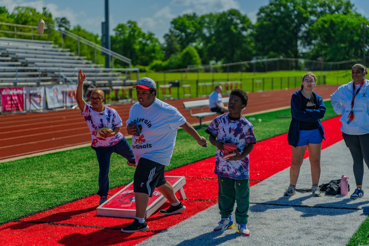 Children compete in a cornhole toss game on a red track at an outdoor school sports event on a sunny day.