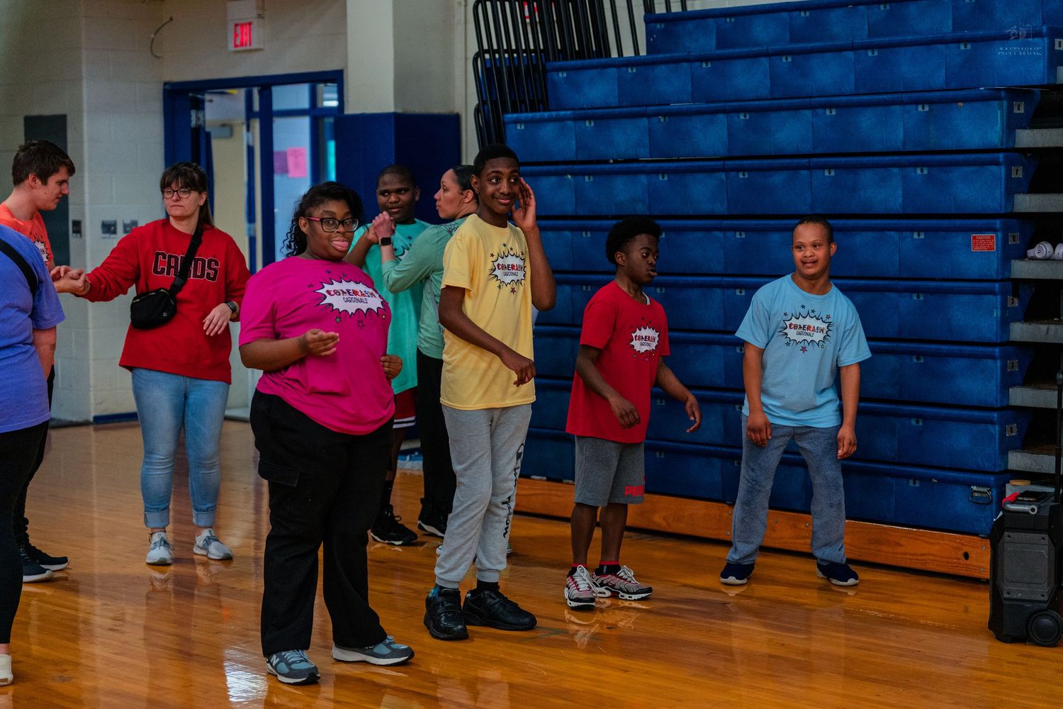 Students dancing and participating in a fun indoor gym activity at school, with blue bleachers in the background.