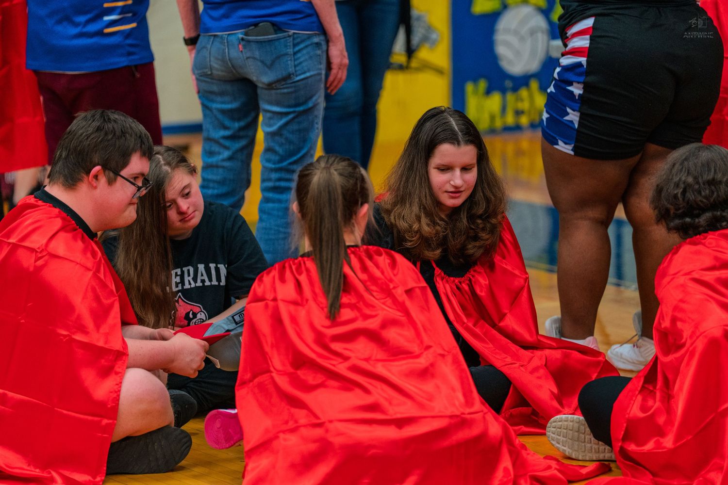 Students in red capes sit together on a gym floor during a school event, engaged in discussion.
