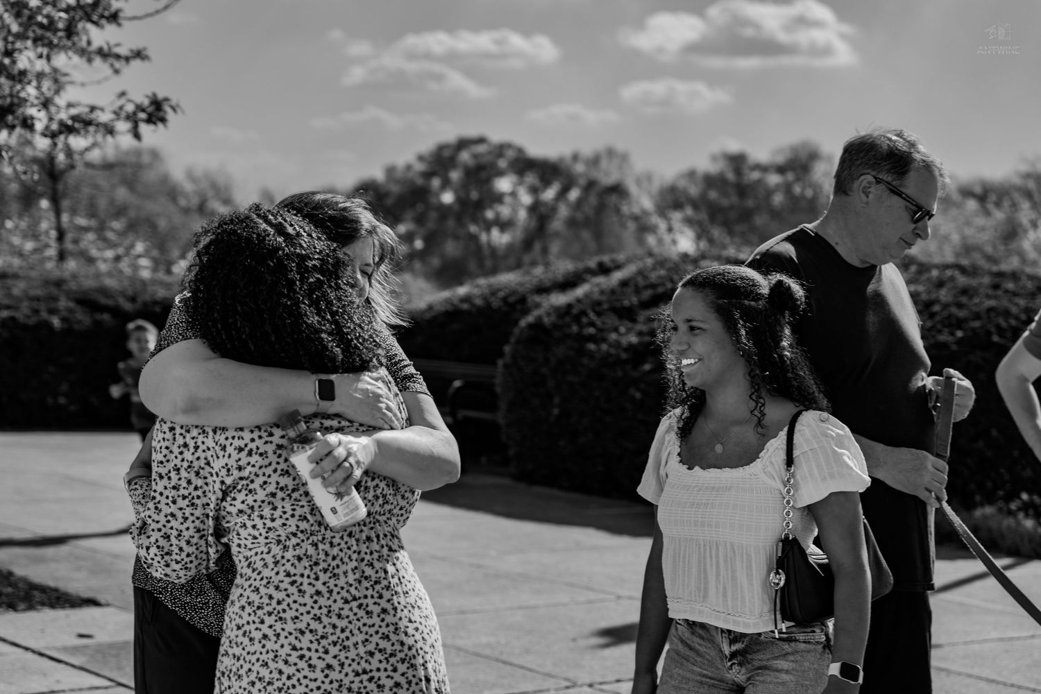 Black and white photo of two women hugging outdoors while another woman smiles nearby.