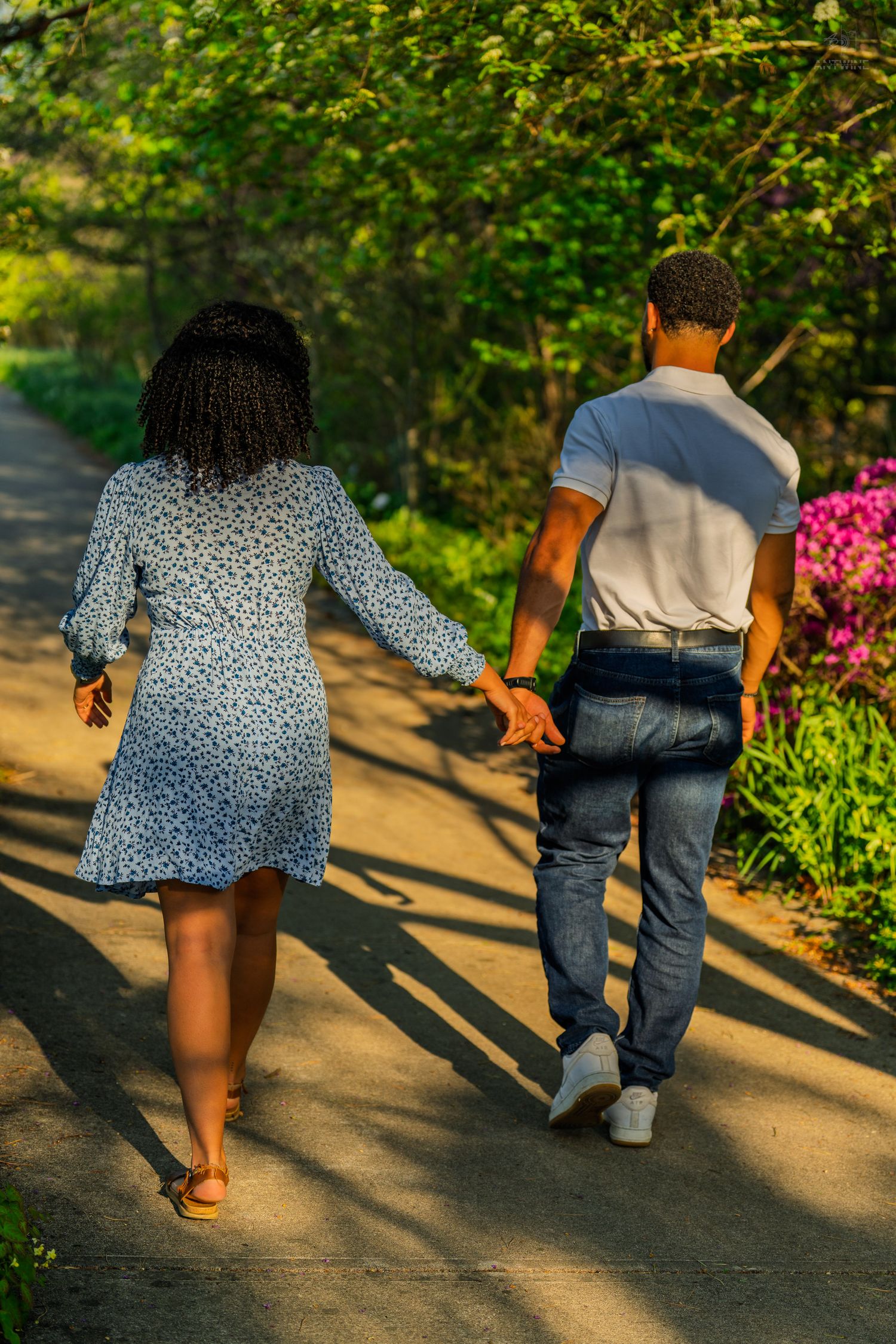 Couple holding hands walking away on a sunlit park path surrounded by lush green trees and flowers.
