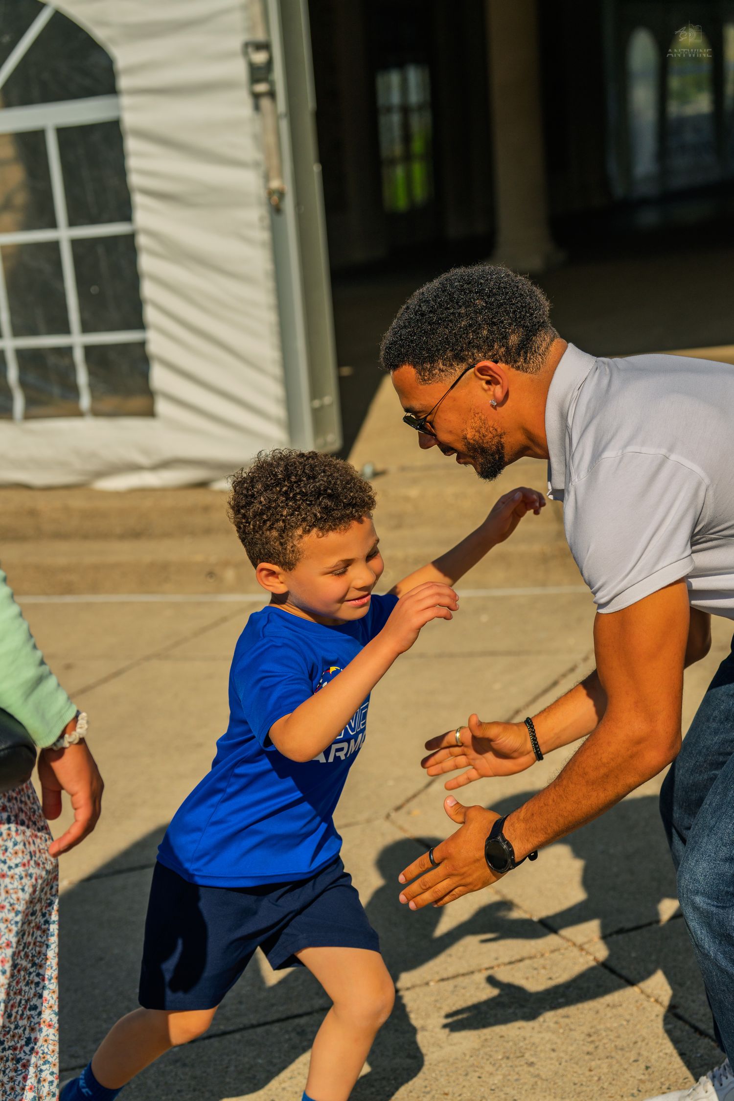 A young boy in a blue shirt joyfully runs to high-five a smiling man outdoors on a sunny day.