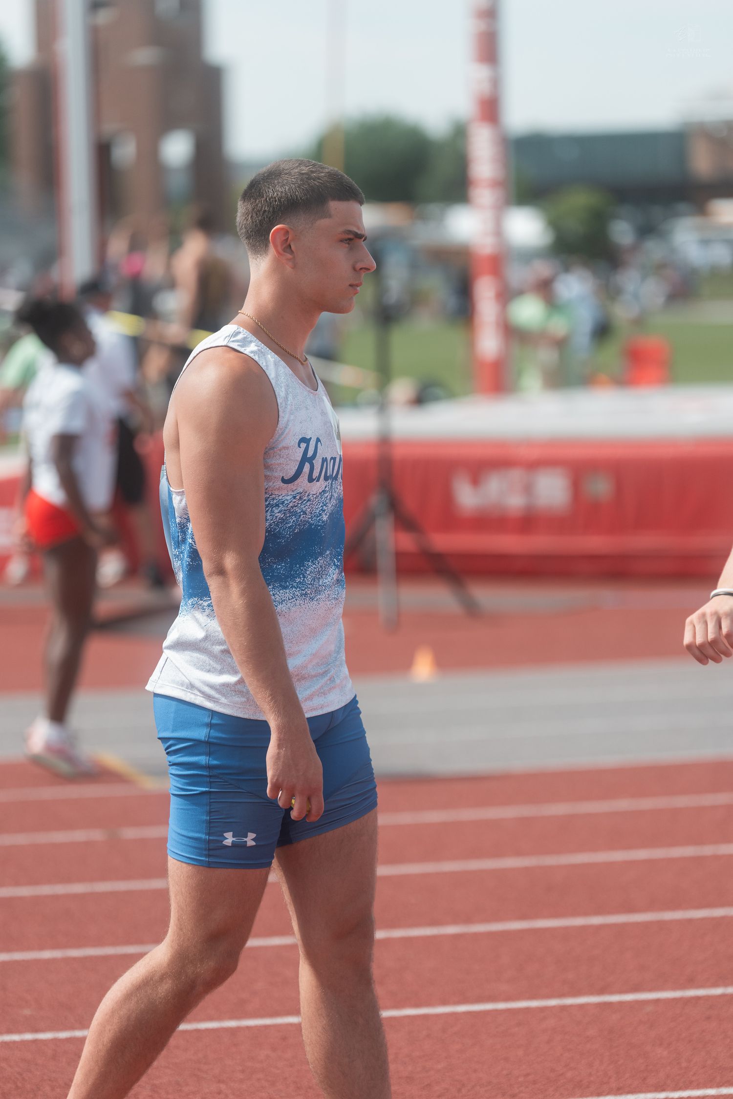 Muscular male athlete in Knights jersey and blue Under Armour shorts stands on outdoor track.