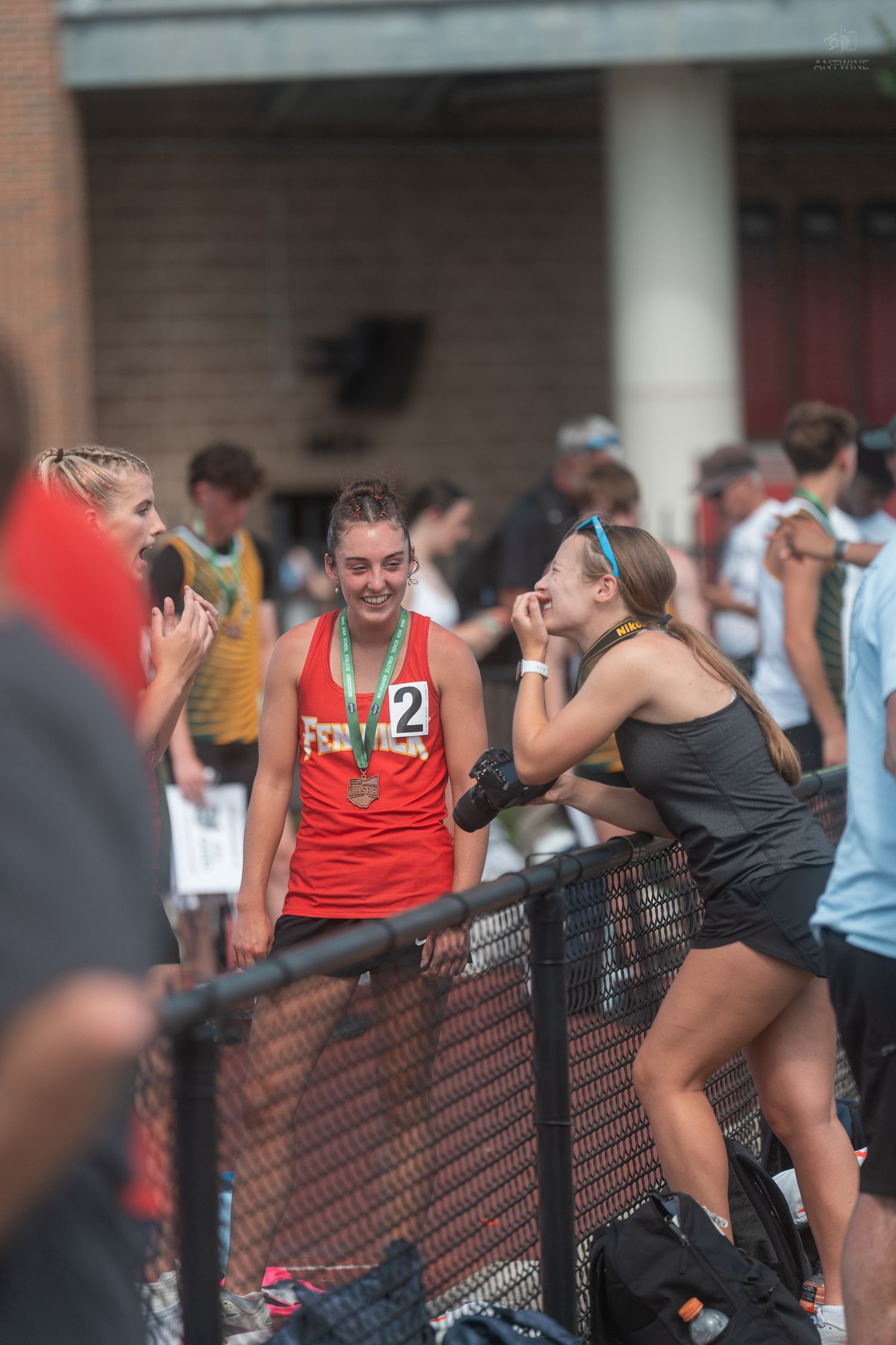 Female track athlete in red number 2 jersey with medal chats with friend at fence.