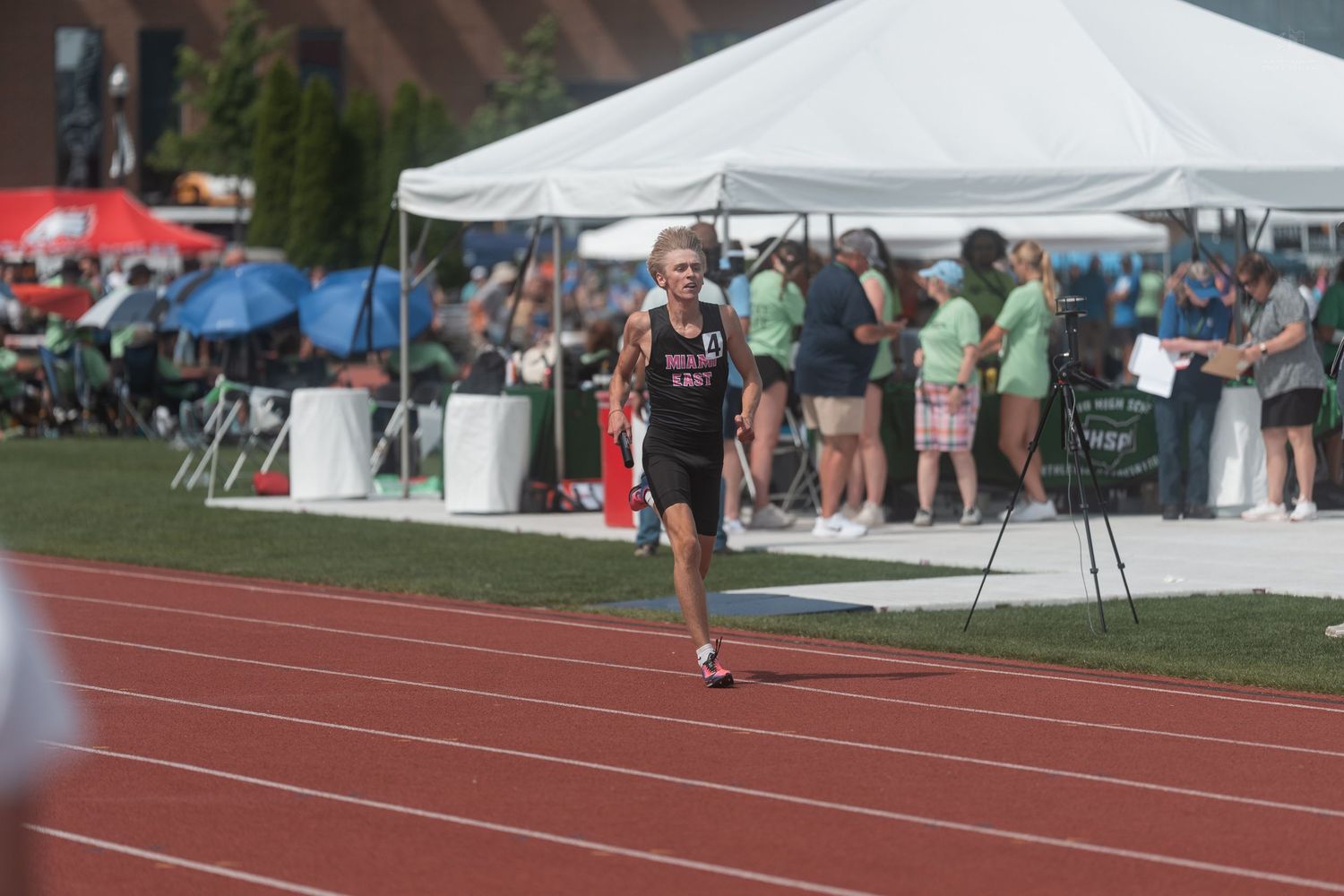 A young male runner in dark uniform races on a red track at a sunny high school track meet.