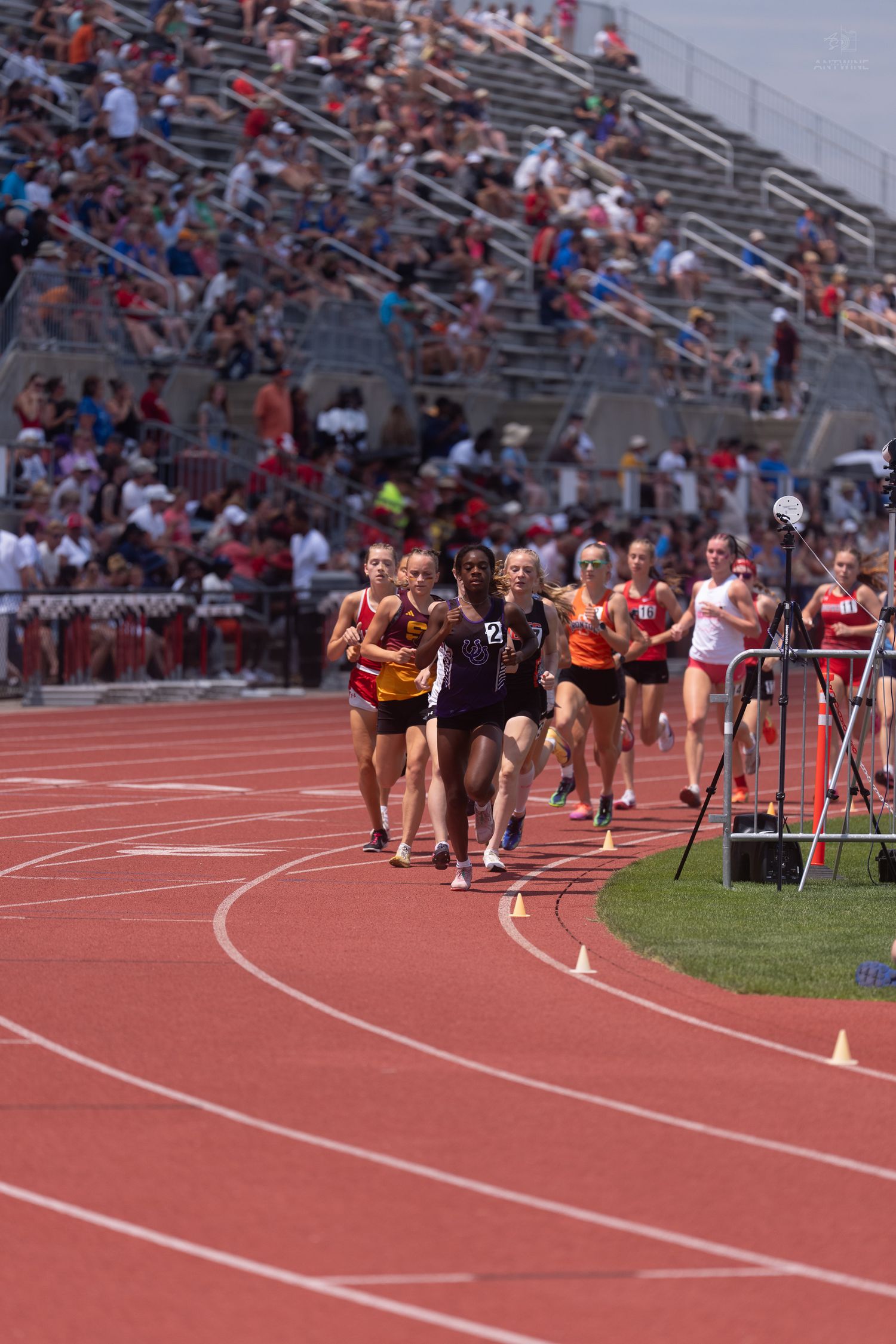 Female athletes race on a red track at a competitive outdoor track and field meet with spectators.