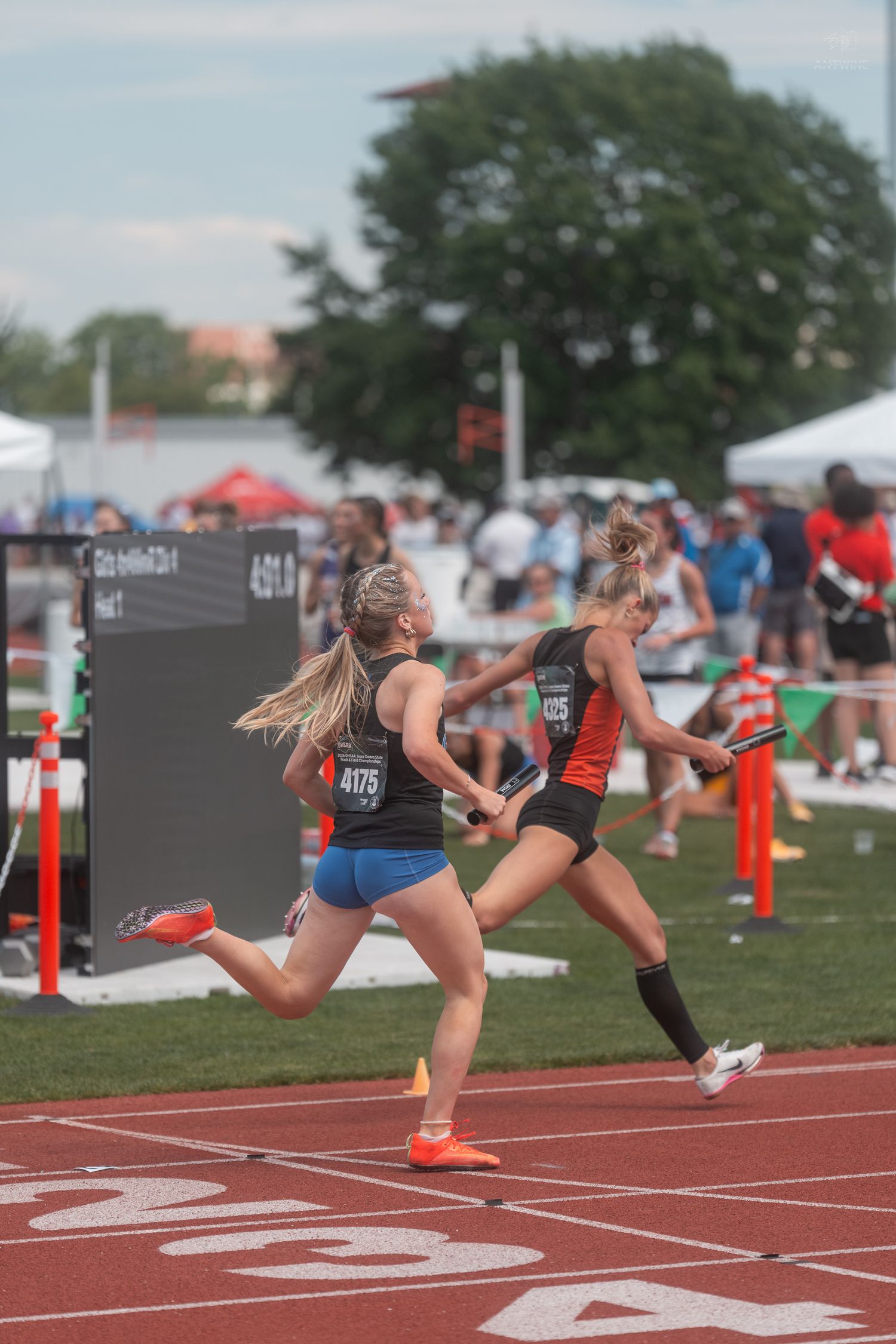 Two female relay runners exchange baton handoff during 4x400m relay at state track championship.