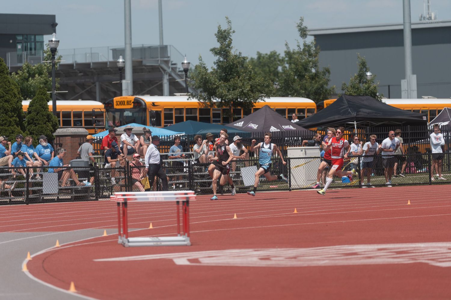 Runners competing in a distance race on a red track with yellow school buses visible in the background.