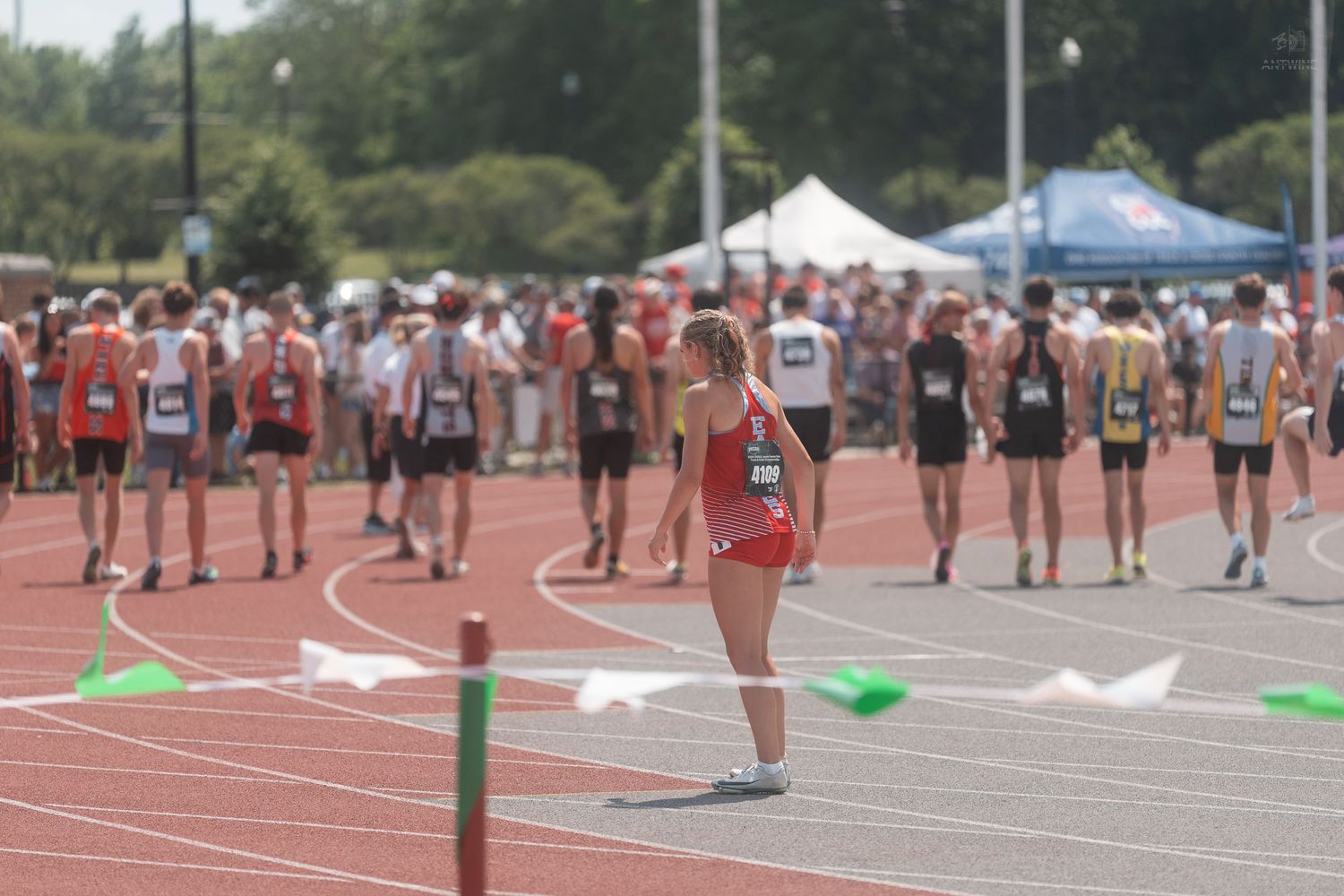 Female athlete stands near hurdle as runners prepare at outdoor track and field meet.