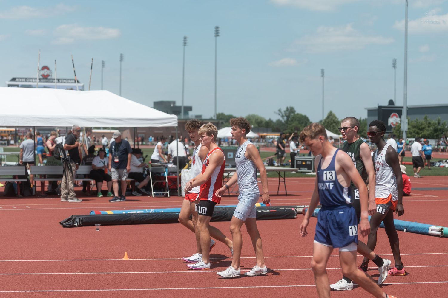 Track and field athletes walk together on a red track at an outdoor stadium during a sunny meet.