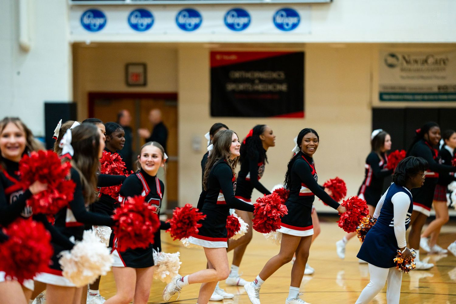 Smiling cheerleaders in red and black uniforms perform with red pom-poms on a basketball court.